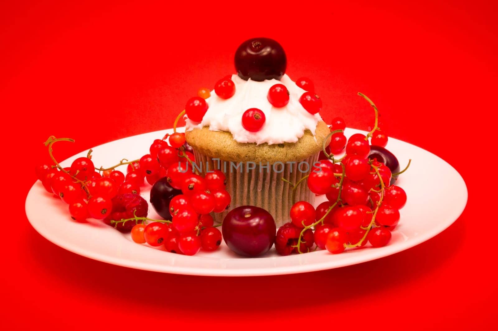White plate with cupcake surrounded by red berries and cherries isolated on red background
