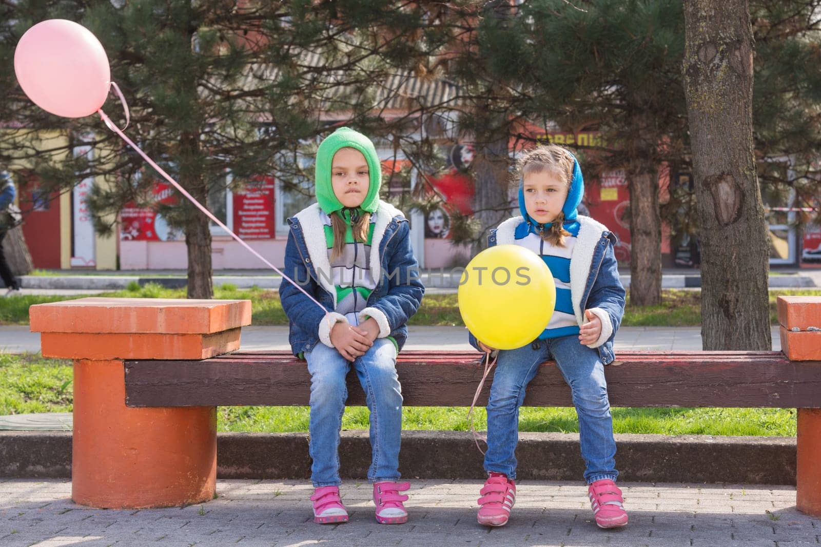 Two sad girl sitting on the bench with balloons in hands