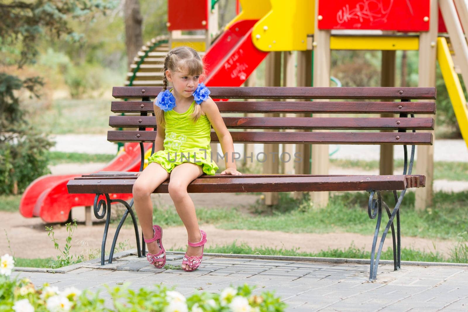 Upset girl bit her lip while sitting on the bench on the background of the playground