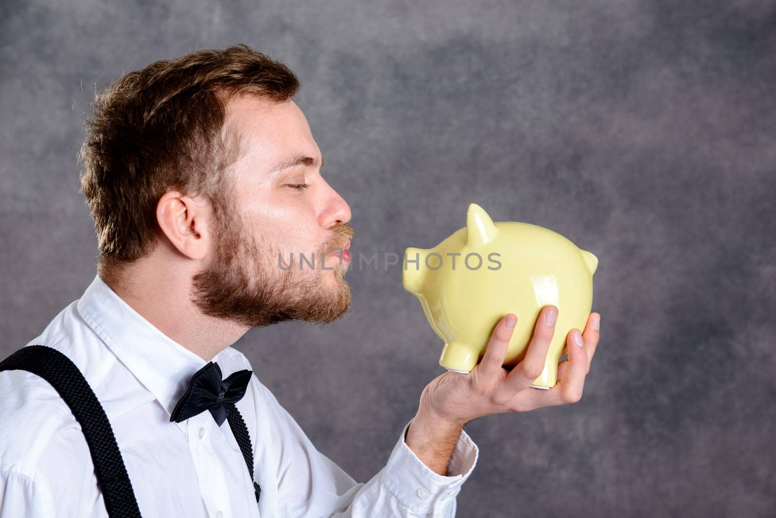 young bearded man in white shirt and bow tie looking at piggy bank