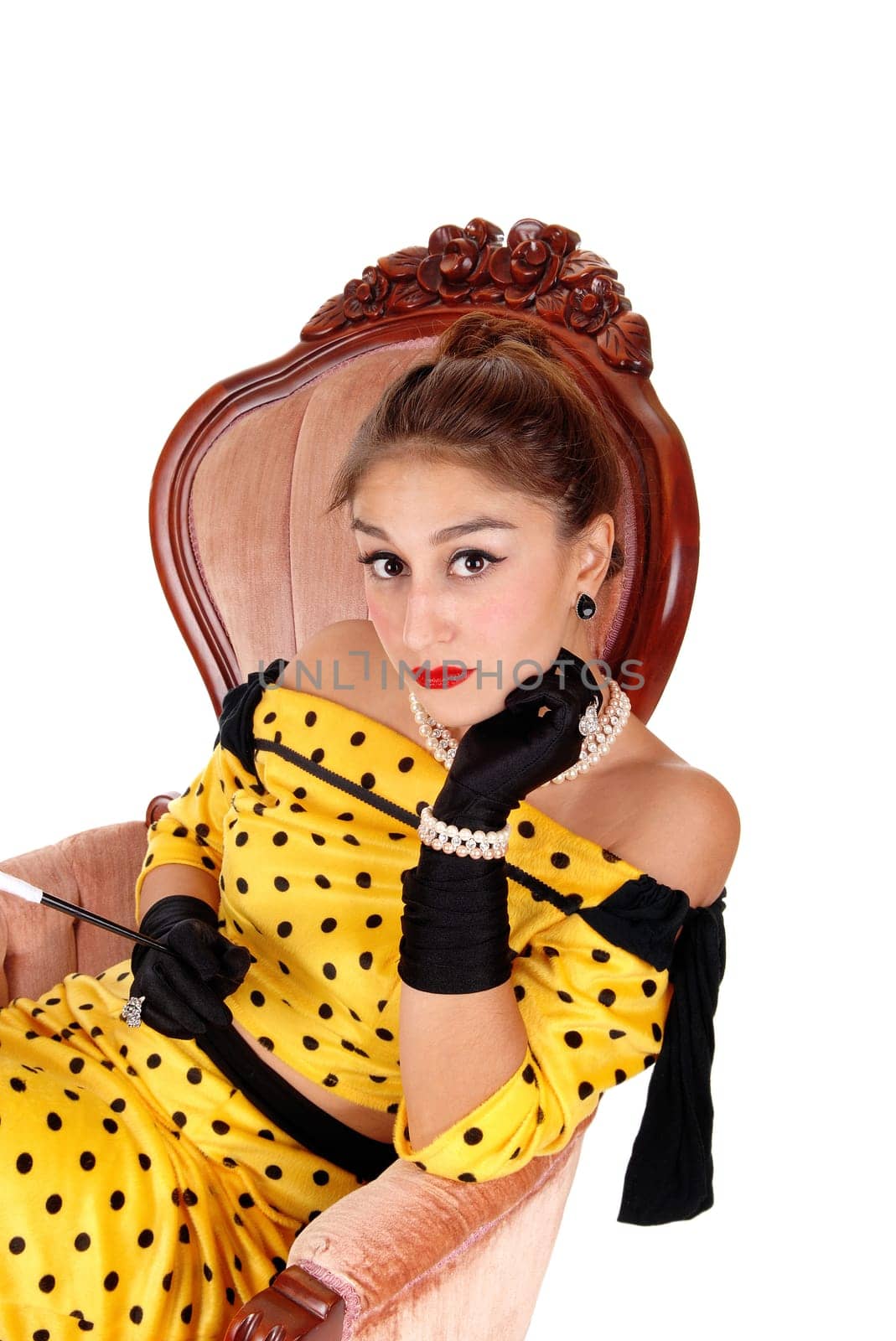A gorgeous young woman sitting in a pink armchair in a yellow dress and.wearing black cloves and jewellery, isolated for white background..