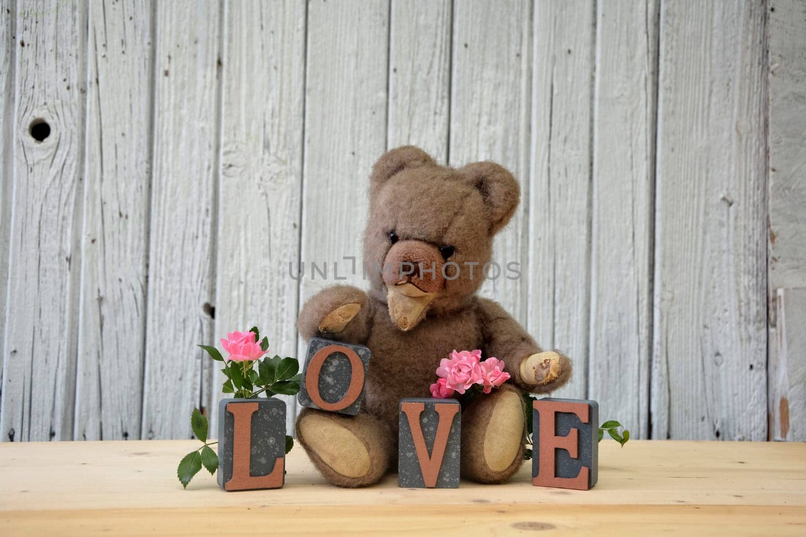 Old antique teddy bear with pink roses and love lettering in front of old white wooden wall