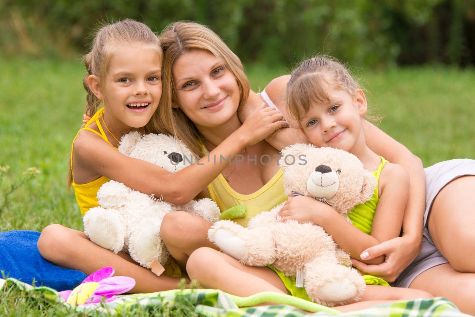 Two daughter kissing and hugging her mother lying on the grass on a picnic