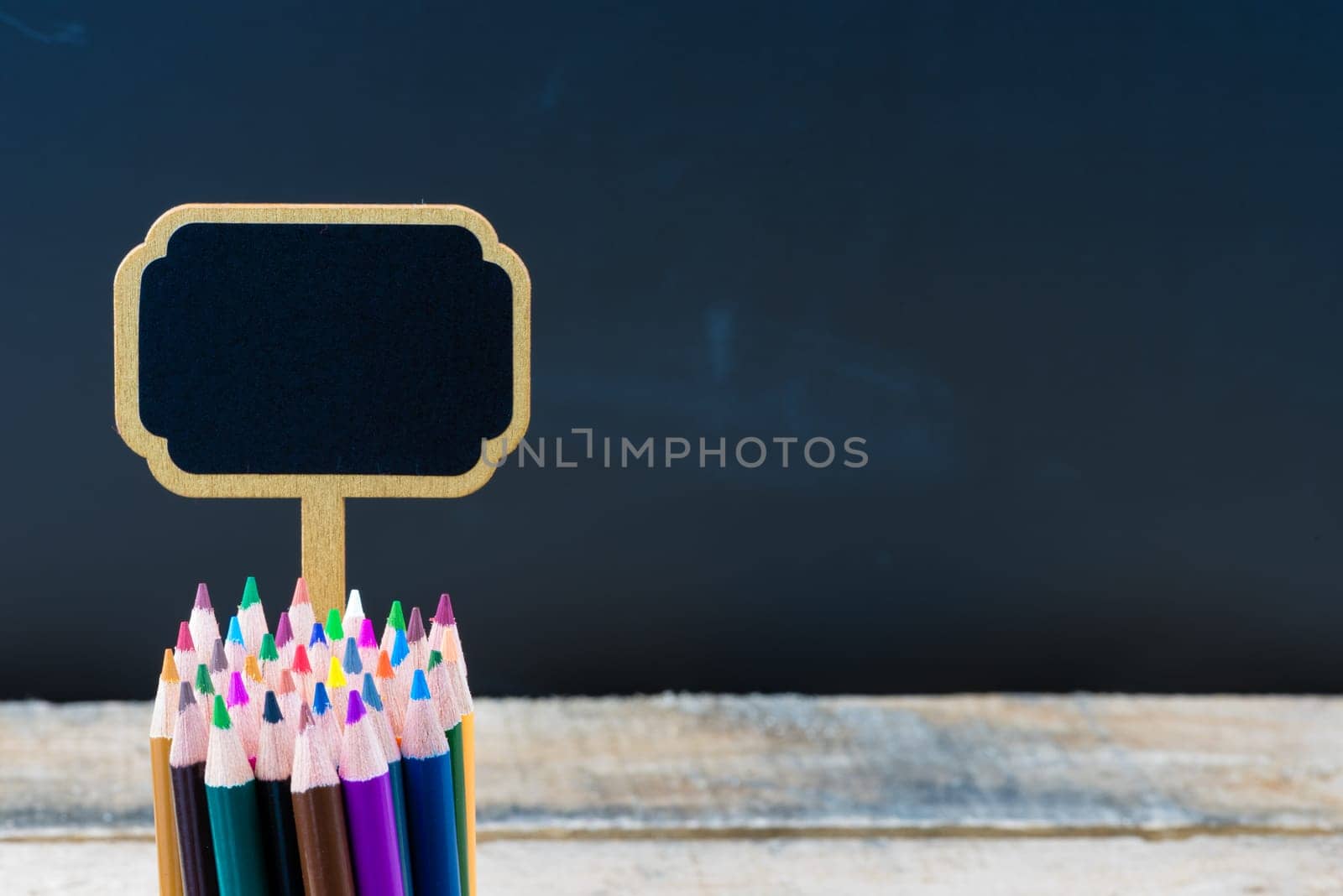 Wooden mini blackboard label and colouring pencils over chalkboard background and table, copy space available, education concept
