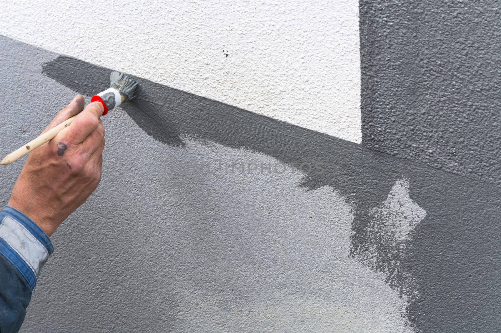 Close-up arm of a painter with paint roll in hand paints a house wall with gray paint.