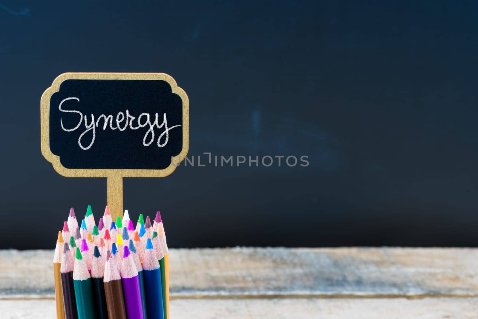 Wooden mini blackboard label with message Synergy and colouring pencils over chalkboard background, copy space available, education concept
