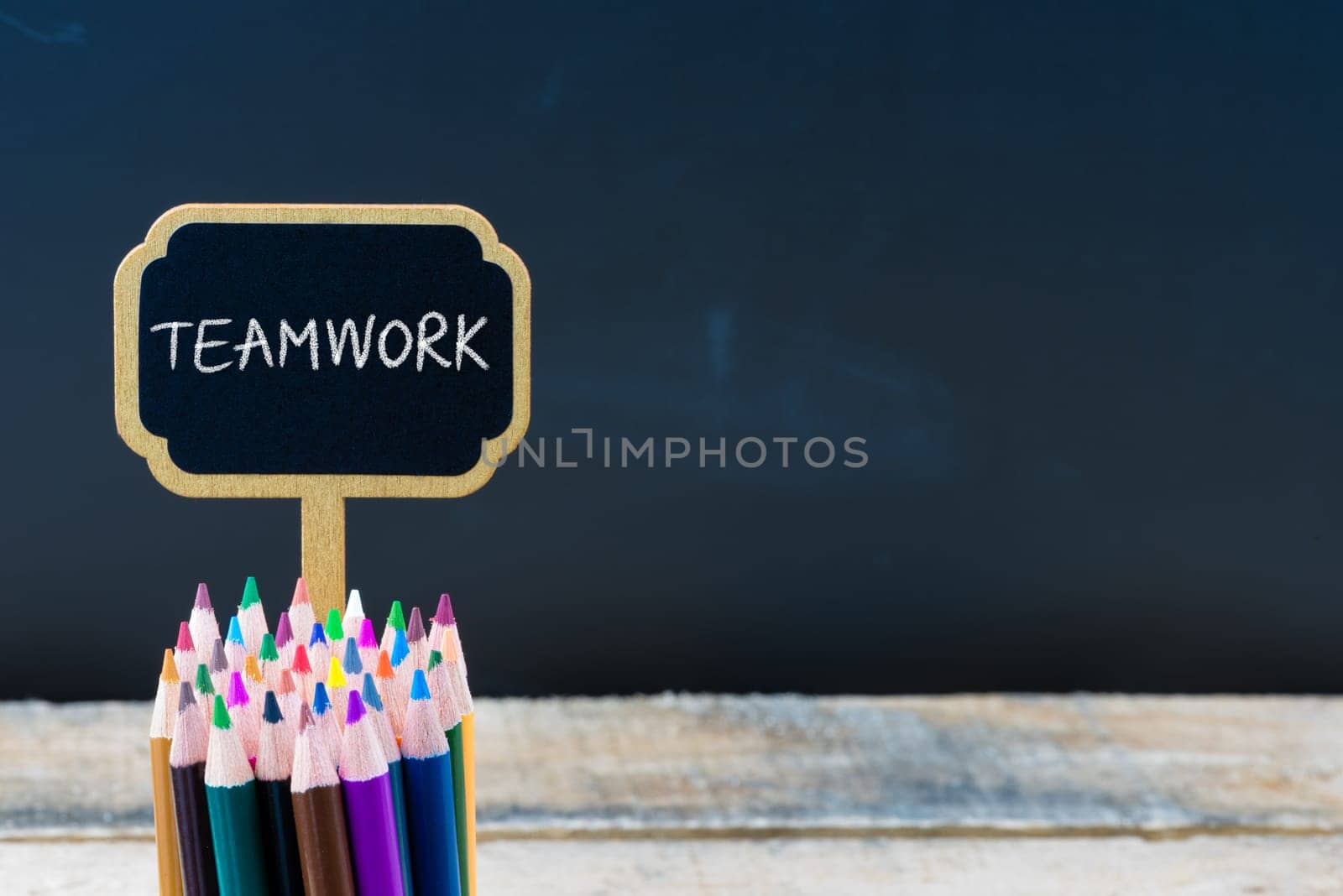 Wooden mini blackboard label with message TEAMWORK and colouring pencils over chalkboard background, copy space available, education concept