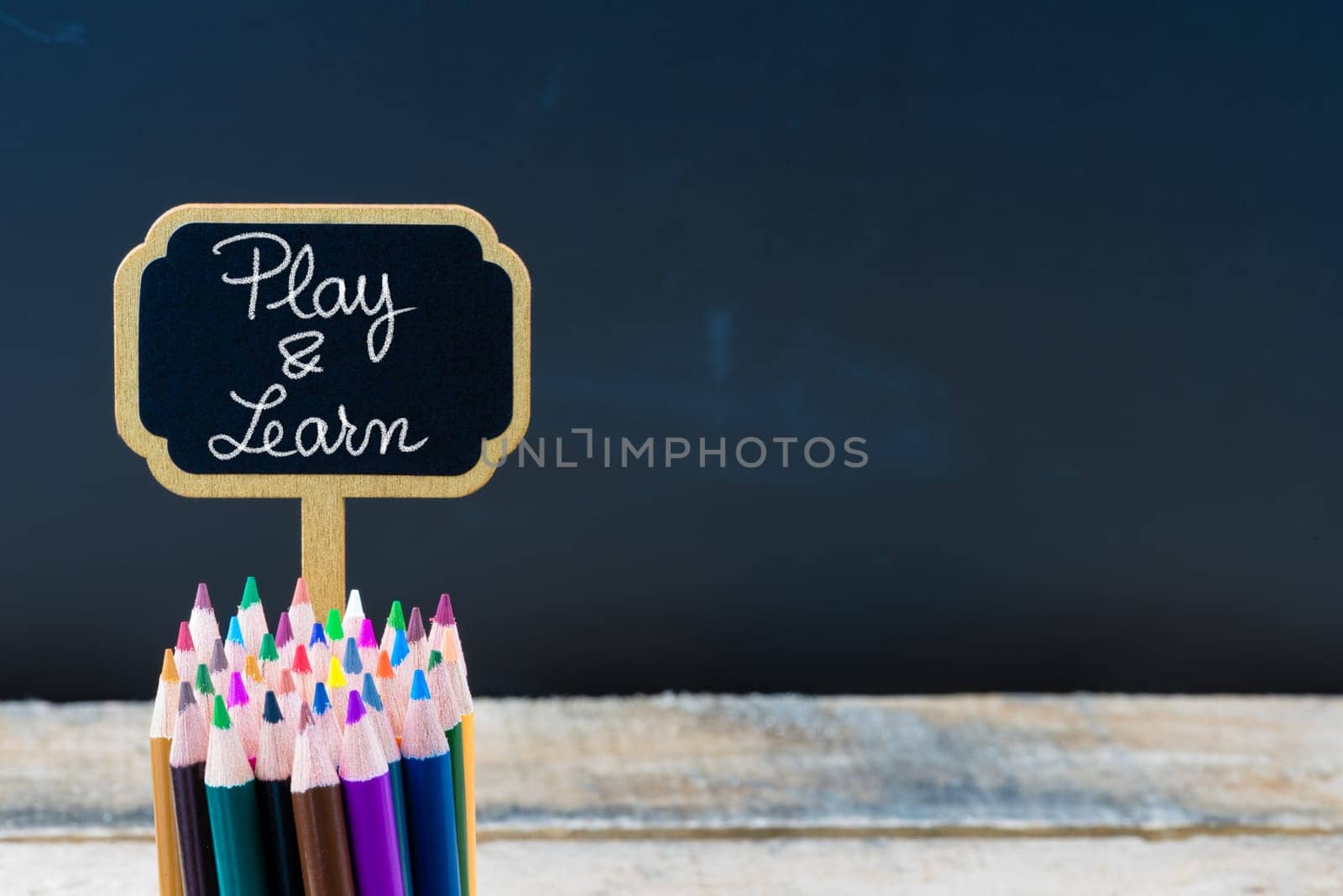 Wooden mini blackboard label with message Play and Learn and colouring pencils over chalkboard background, copy space available, education concept