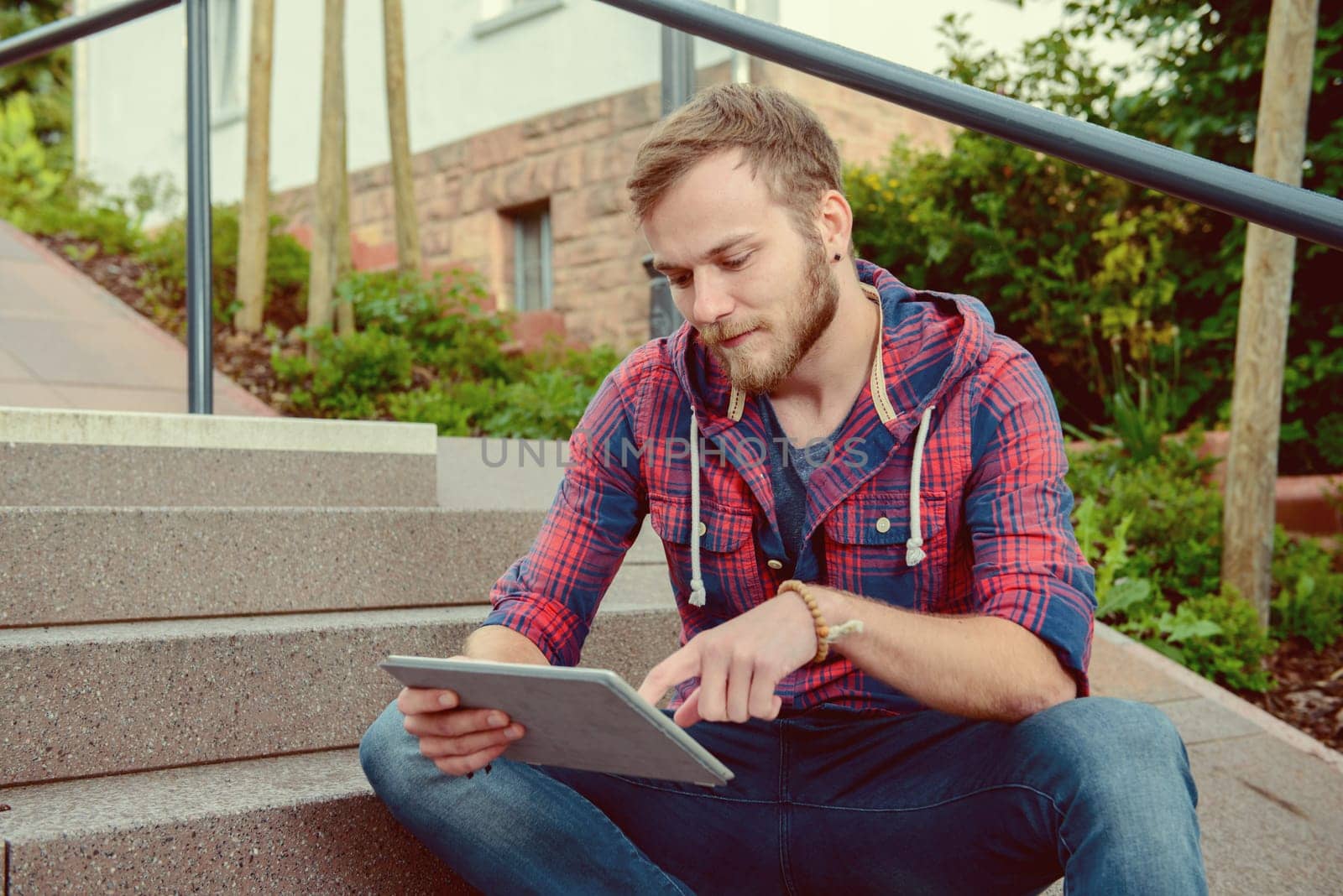 young man in checkered shirt sitting on stairs and using tablet PC