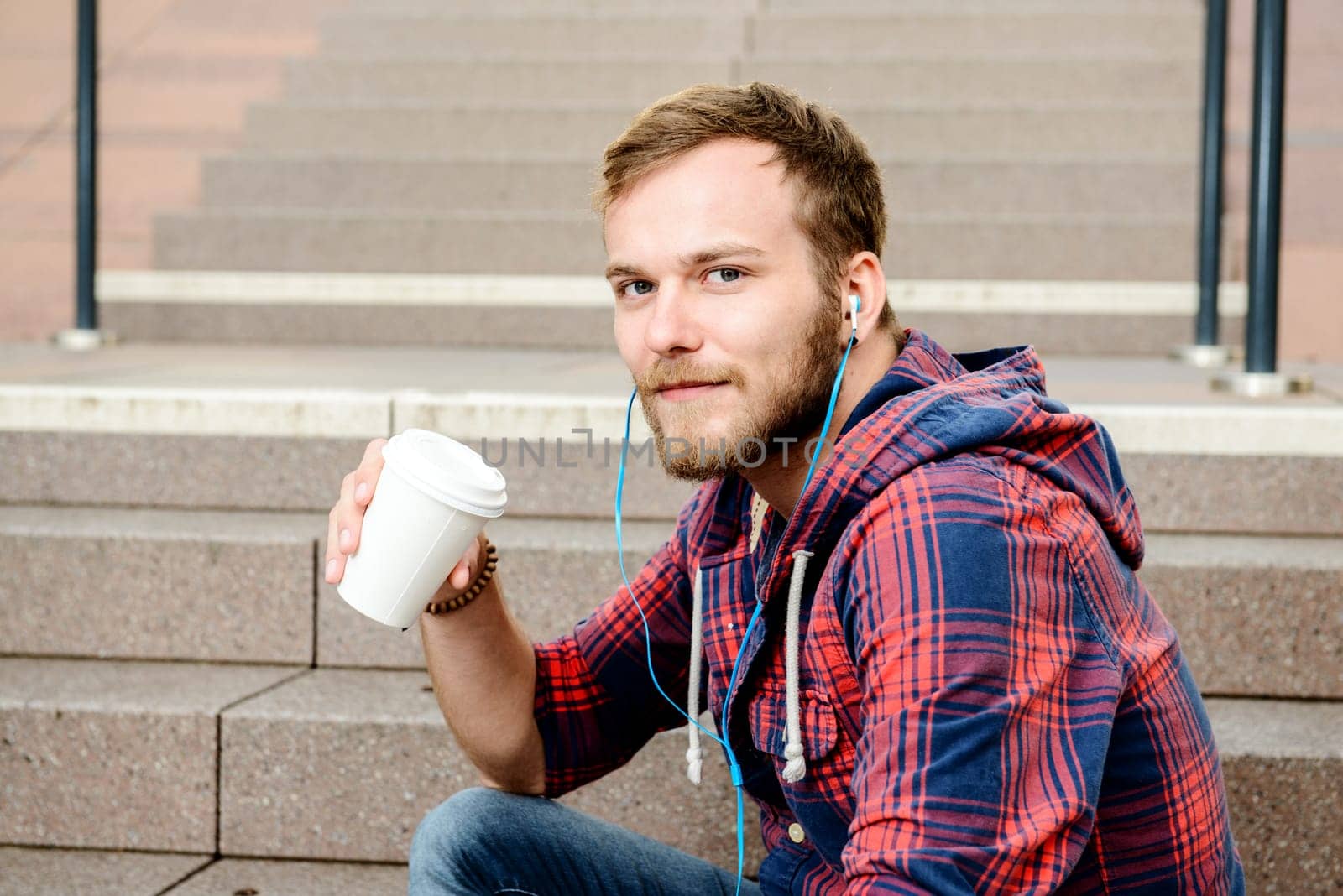 young bearded man in checkered shirt sitting on stairs listening music and drinking coffee