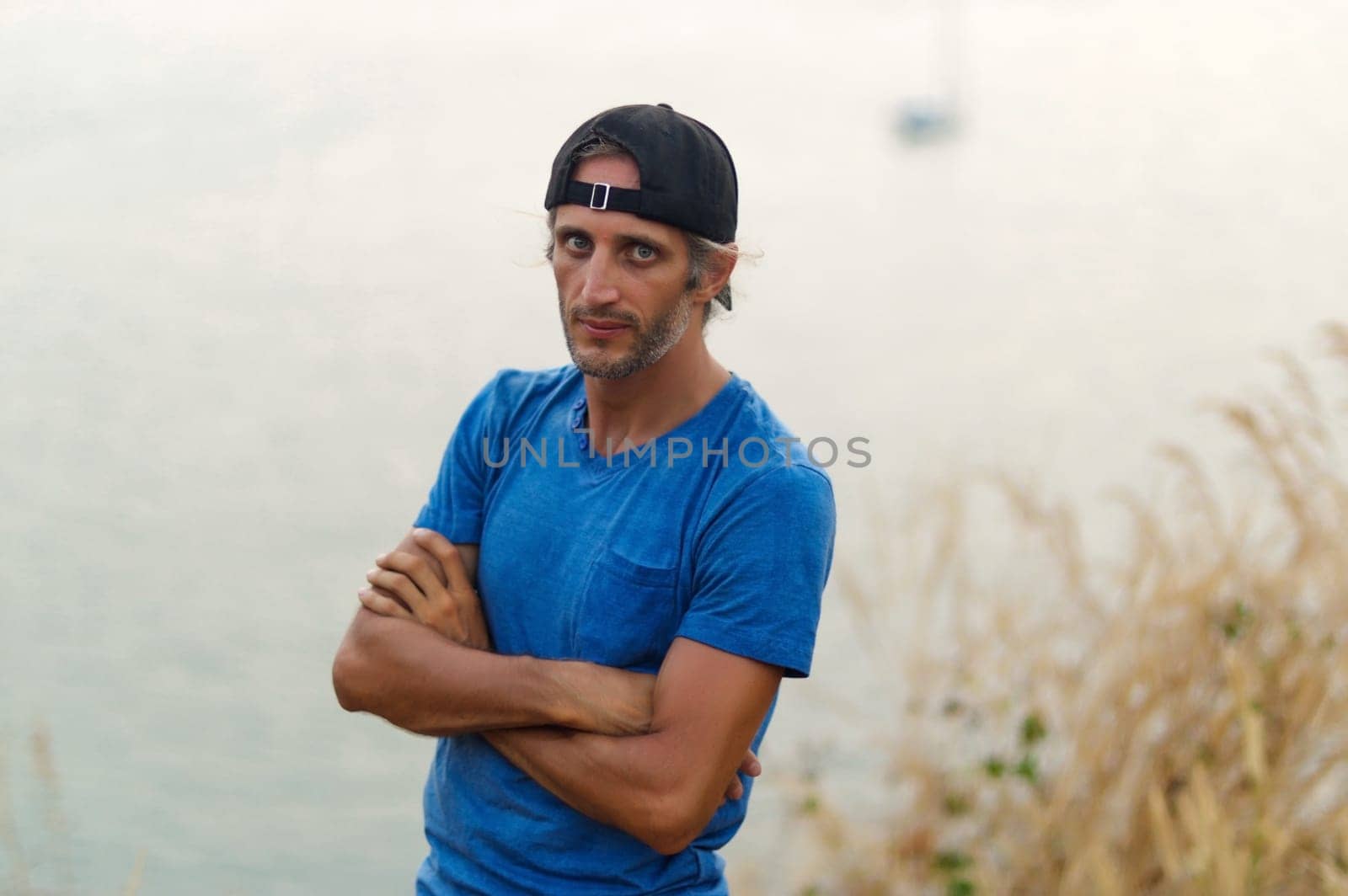 a happy Young Bearded Man sitting on the stone railings in city park wearing in blank blue t-shirt and black Cap