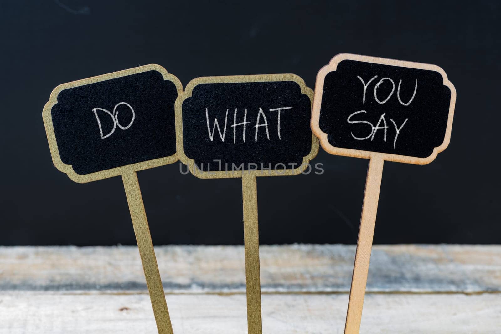 Business message DO WHAT YOU SAY written with chalk on wooden mini blackboard labels, defocused chalkboard and wood table in background