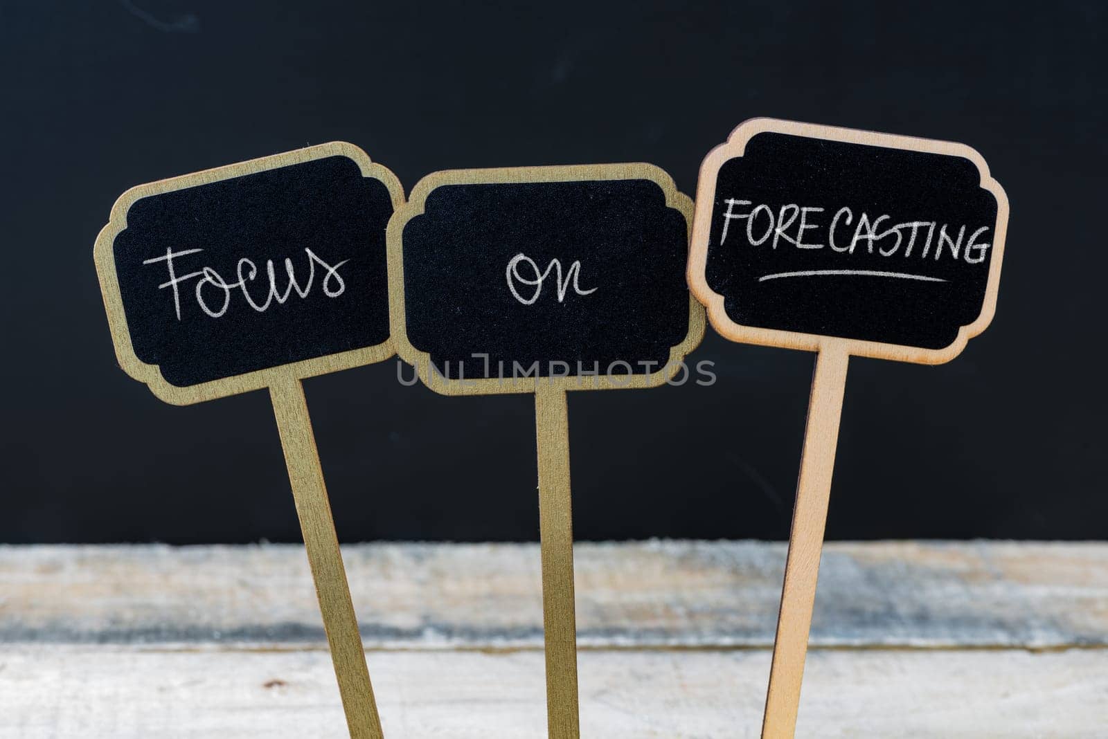 Business message FOCUS ON FORECASTING written with chalk on wooden mini blackboard labels, defocused chalkboard and wood table in background
