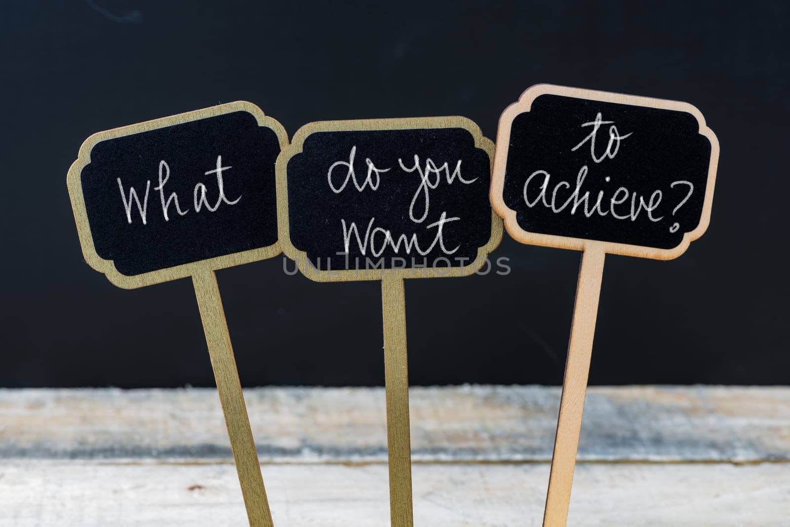 Business message WHAT DO YOU WANT TO ACHIEVE ? written with chalk on wooden mini blackboard labels, defocused chalkboard and wood table in background