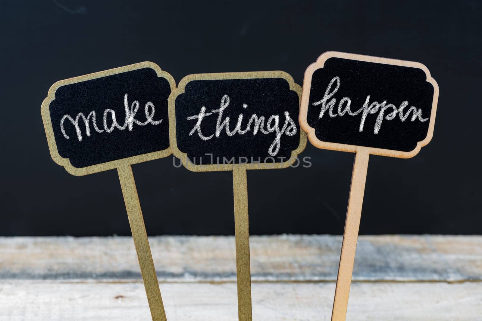 Business message MAKE THINGS HAPPEN written with chalk on wooden mini blackboard labels, defocused chalkboard and wood table in background