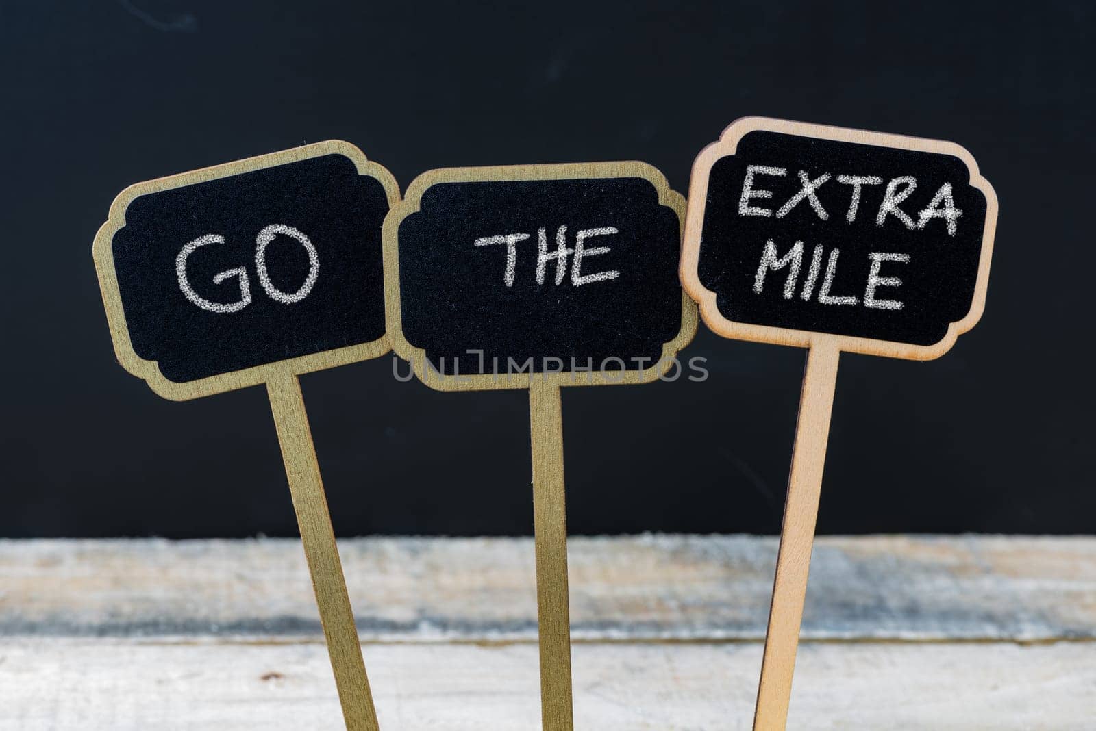 Business message GO THE EXTRA MILE written with chalk on wooden mini blackboard labels, defocused chalkboard and wood table in background