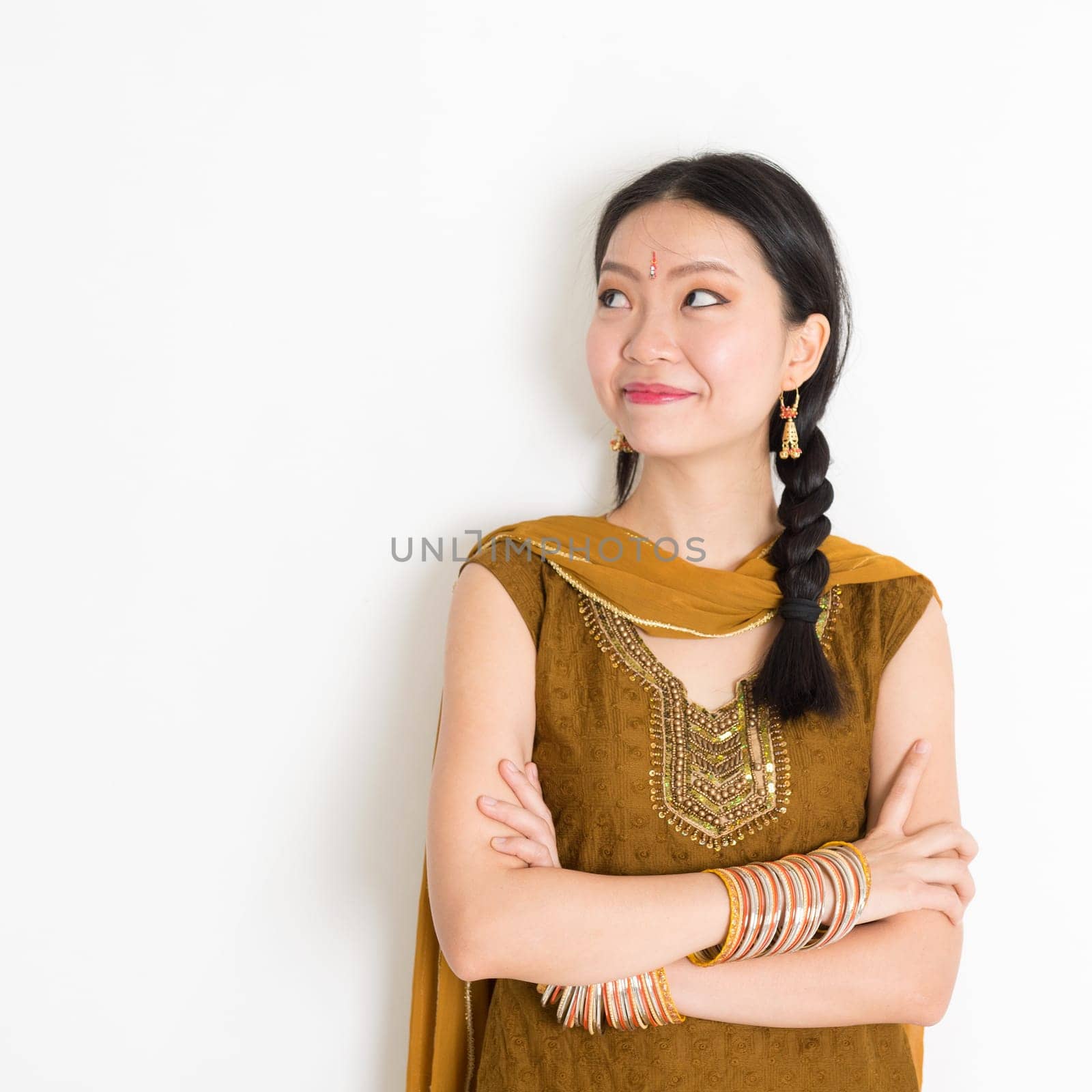 Portrait of arms crossed mixed race Indian Chinese girl in traditional Punjabi dress looking side upward, standing on plain white background.