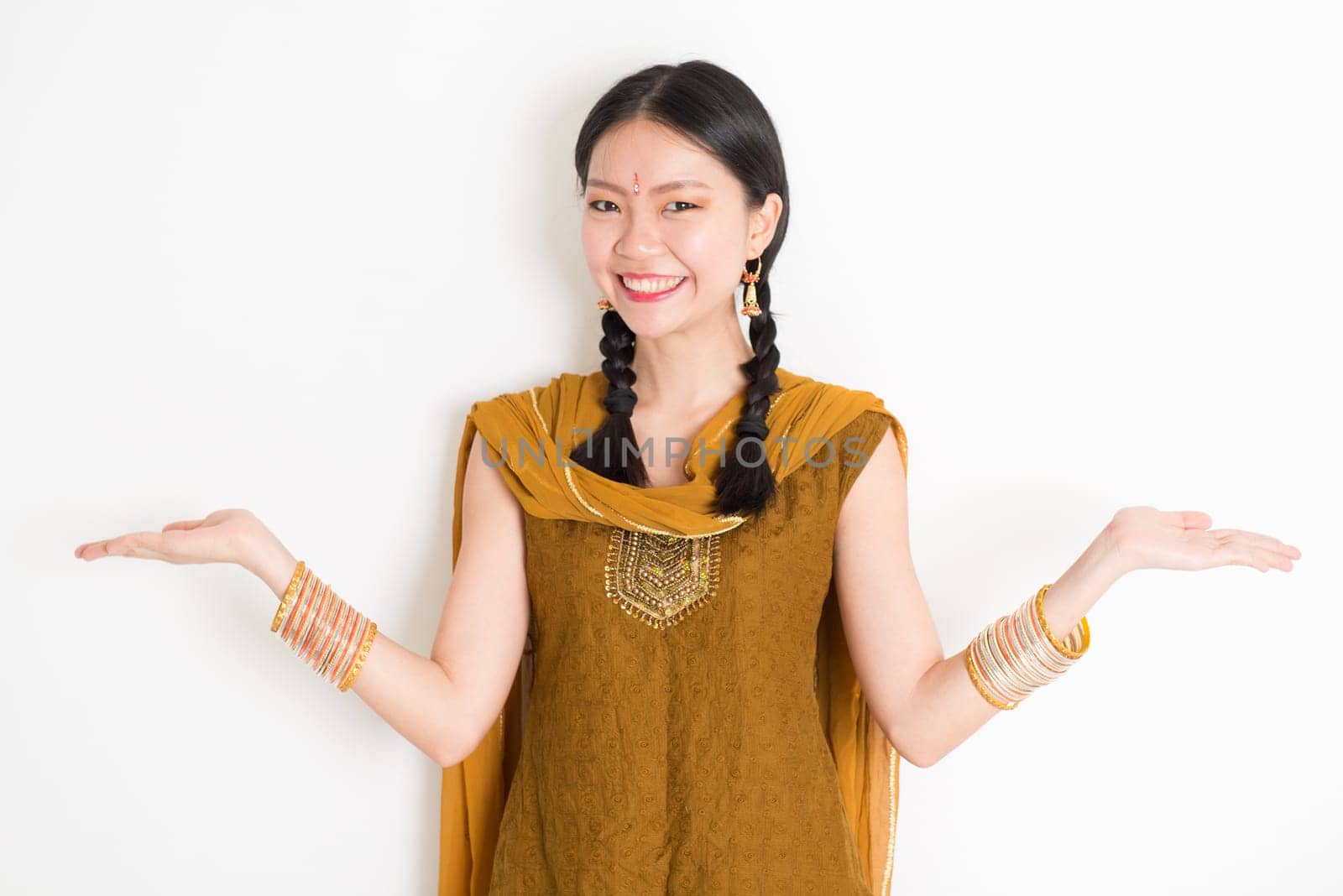 Portrait of young mixed race Indian Chinese woman in traditional punjabi dress hands showing somethings, standing on plain white background.