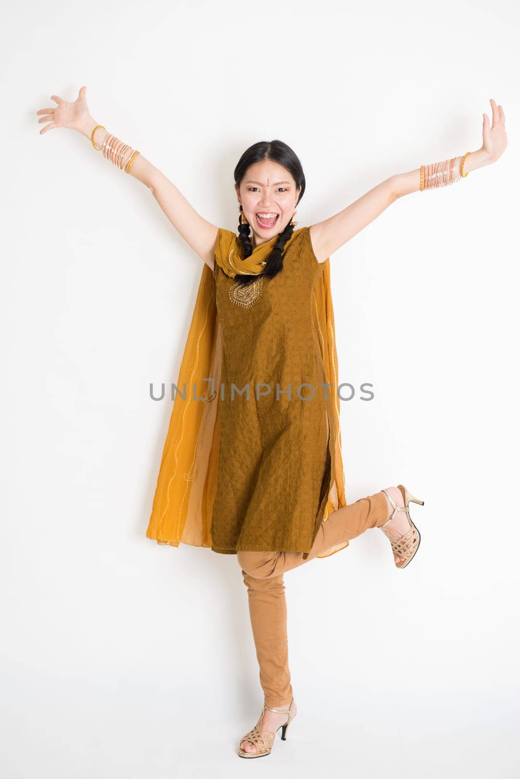 Portrait of excited mixed race Indian Chinese girl in traditional punjabi dress arms raised, full length standing on plain white background.