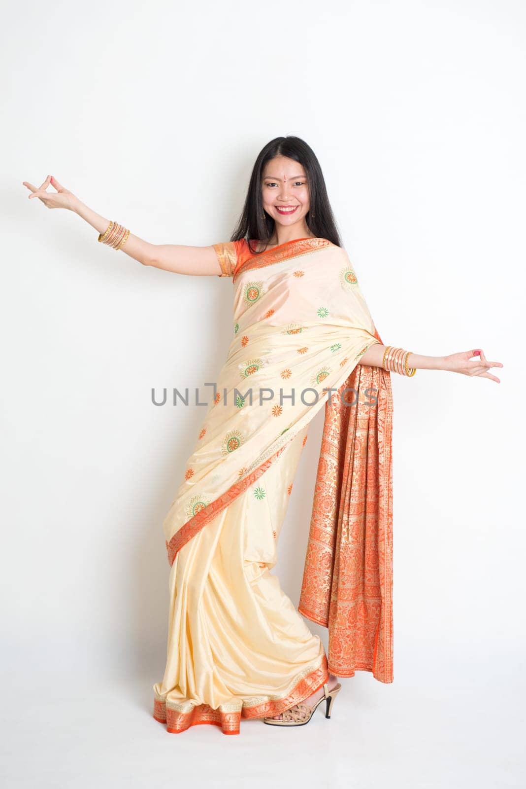 Full length portrait of young mixed race Indian Chinese woman in traditional sari dress dancing, on plain background.