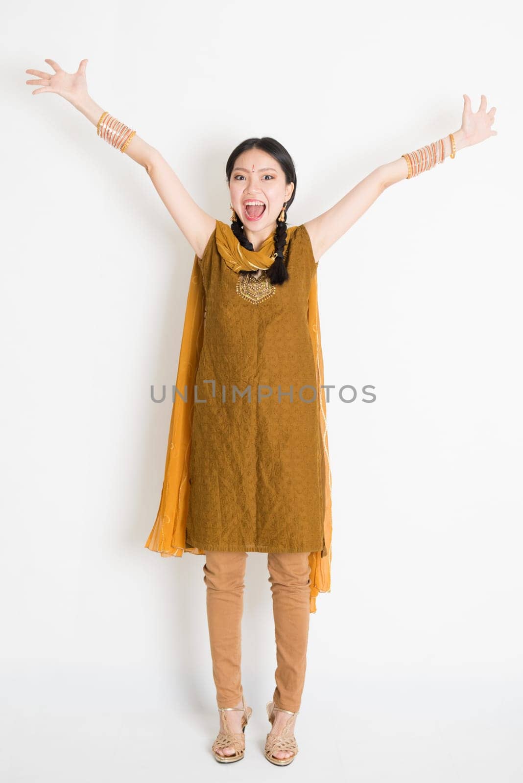 Portrait of excited mixed race Indian Chinese woman in traditional punjabi dress arms outstretched, full length standing on plain white background.
