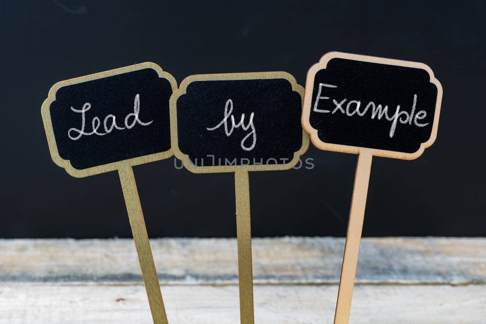 Business message Lead By Example written with chalk on wooden mini blackboard labels, defocused chalkboard and wood table in background