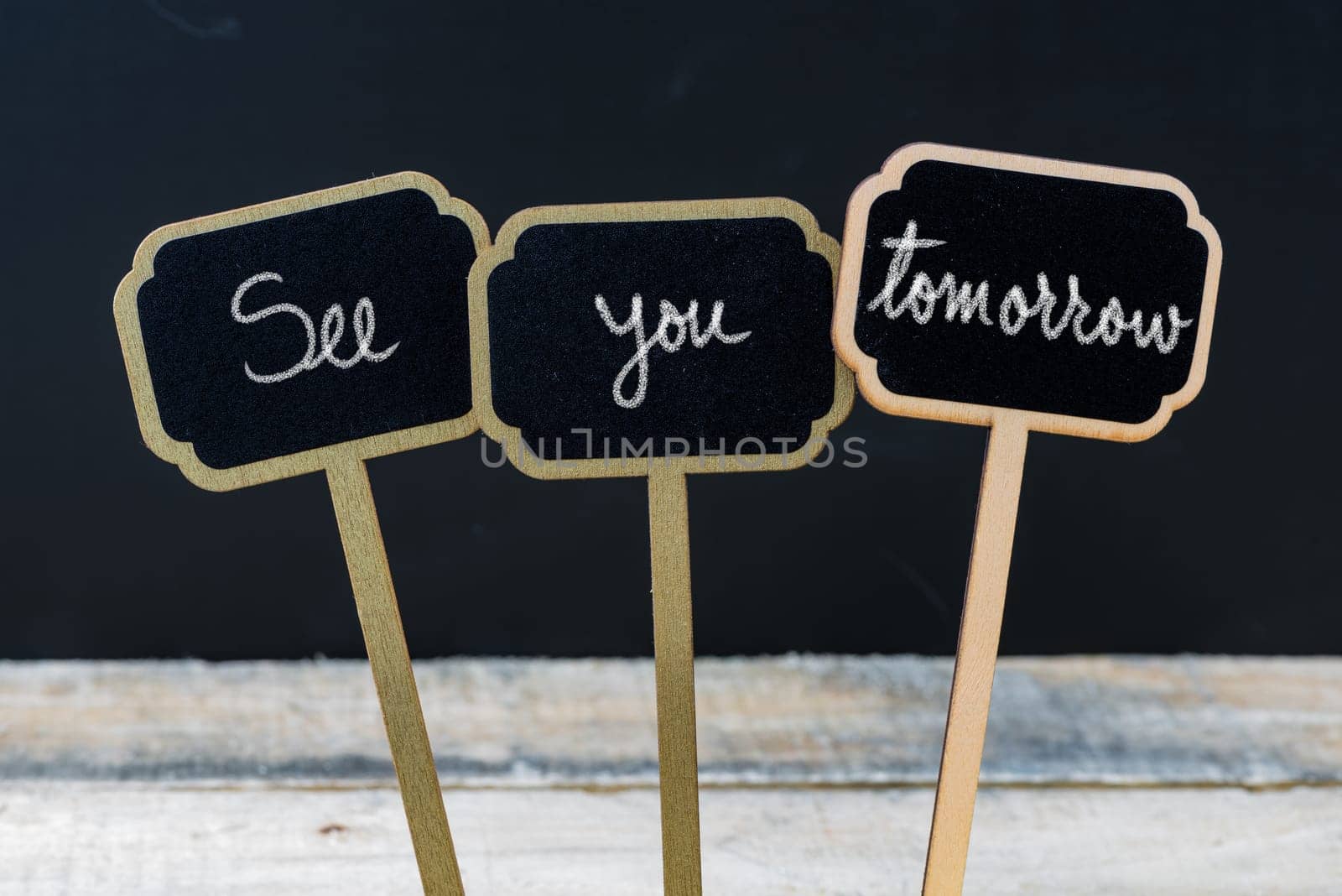 Business message See You Tomorrow written with chalk on wooden mini blackboard labels, defocused chalkboard and wood table in background