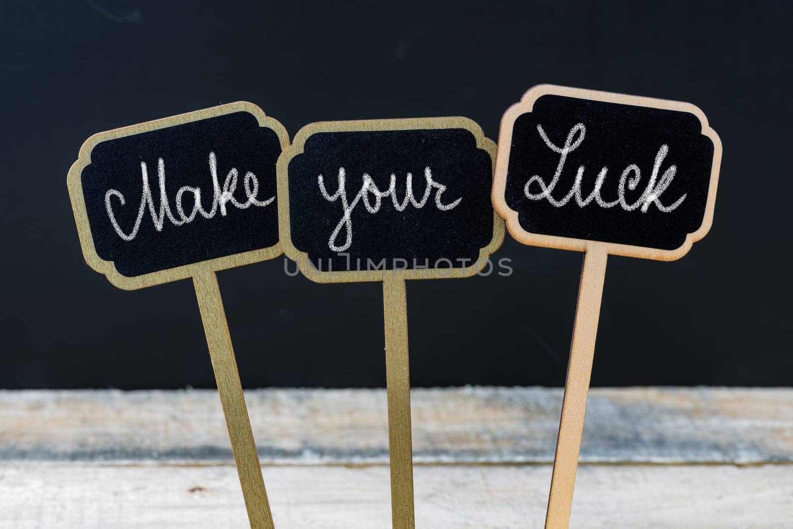 Motivational message Make Your Luck written with chalk on wooden mini blackboard labels, defocused chalkboard and wood table in background