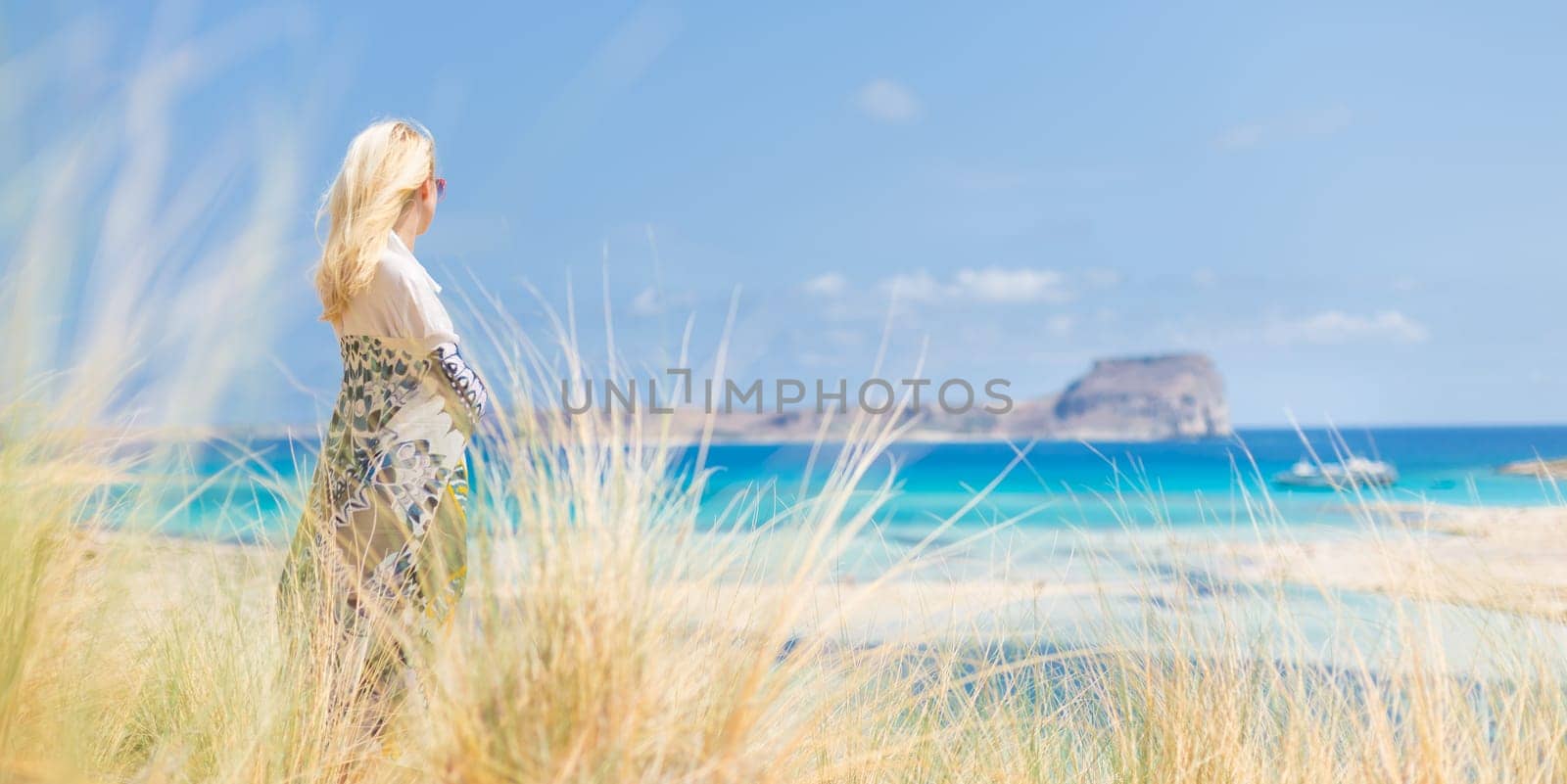 Relaxed woman wrapped in colorful scarf, enjoying sun, freedom and life at beautiful Balos beach in Greece. Concept of holidays, vacations, freedom, joy and well being.