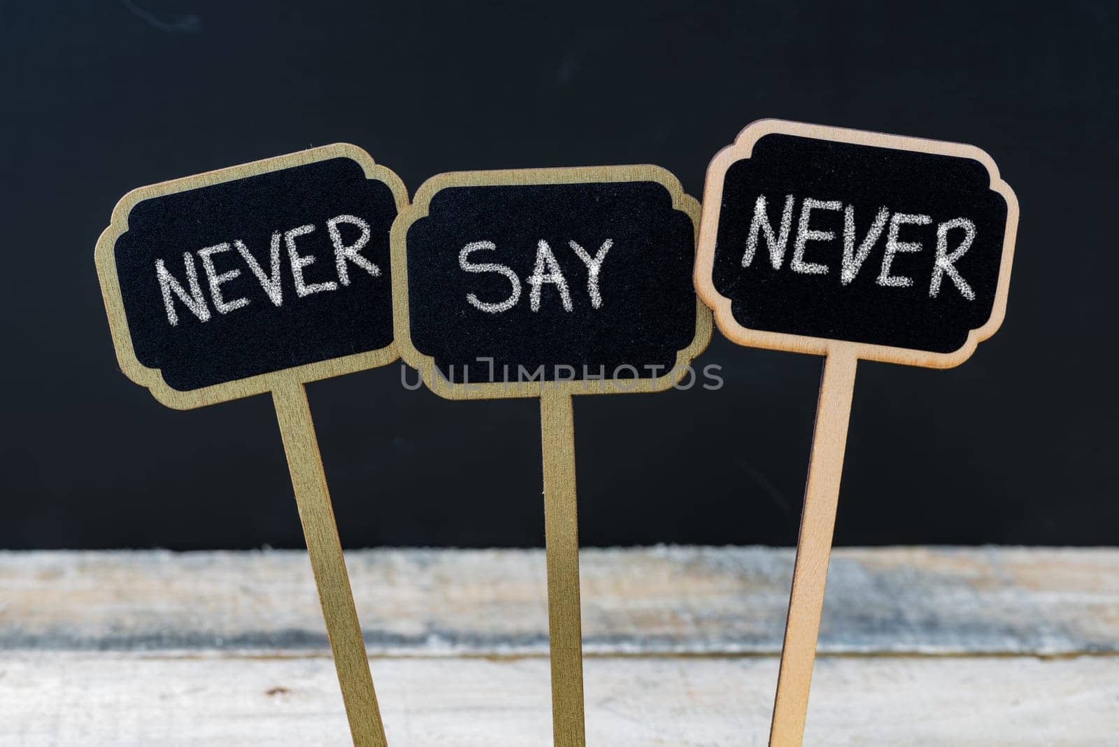 Motivational message NEVER SAY NEVER written with chalk on wooden mini blackboard labels, defocused chalkboard and wood table in background