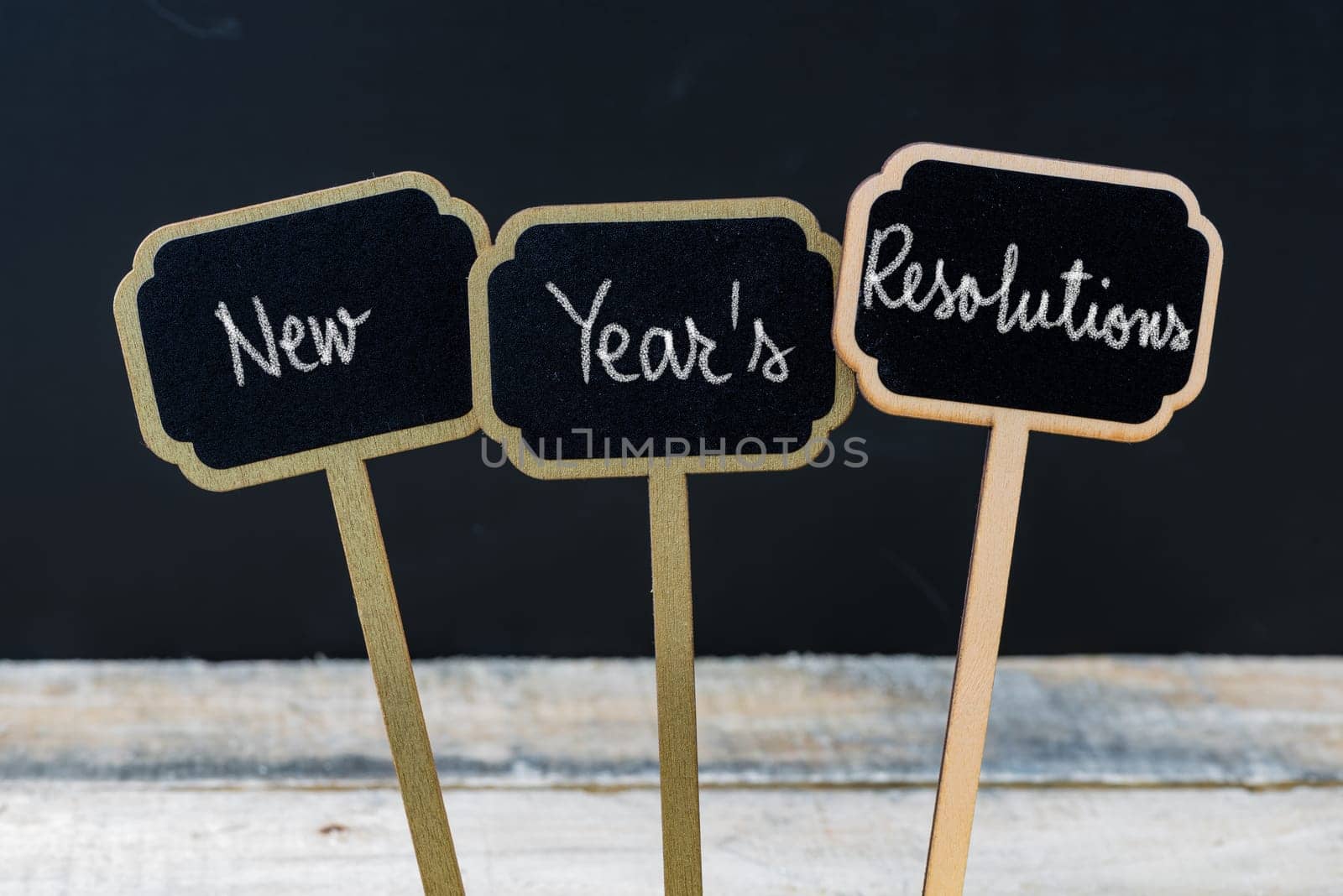 Motivational message New Years Resolutions written with chalk on wooden mini blackboard labels, defocused chalkboard and wood table in background
