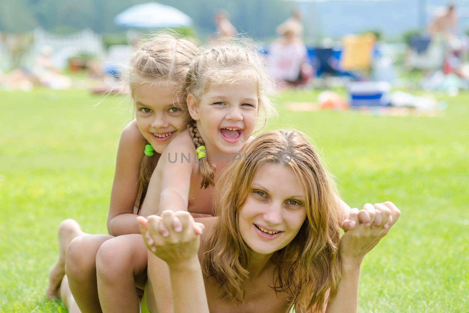 Two daughters are sitting on the mother's back, hot summer day relaxing on the green lawn beautiful beaches