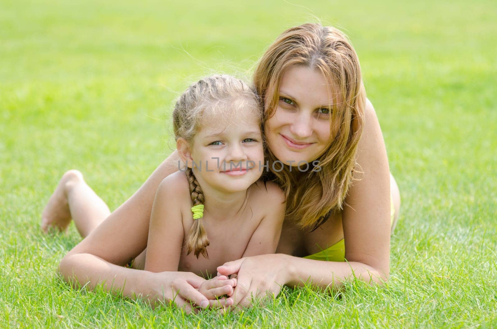 Young mother and five year old daughter hugging lying on green grass and looking to the frame