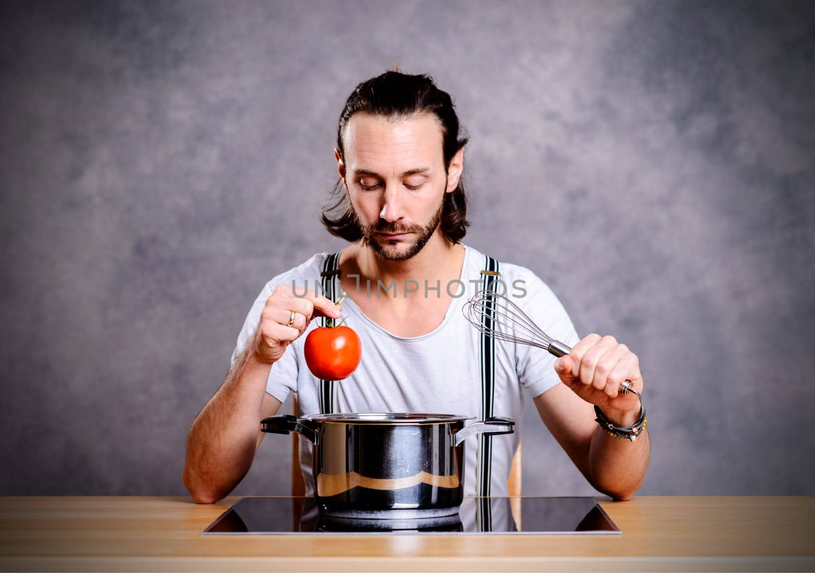 young bearded man with cooking pot, eggwhish and tomato in front of gray background