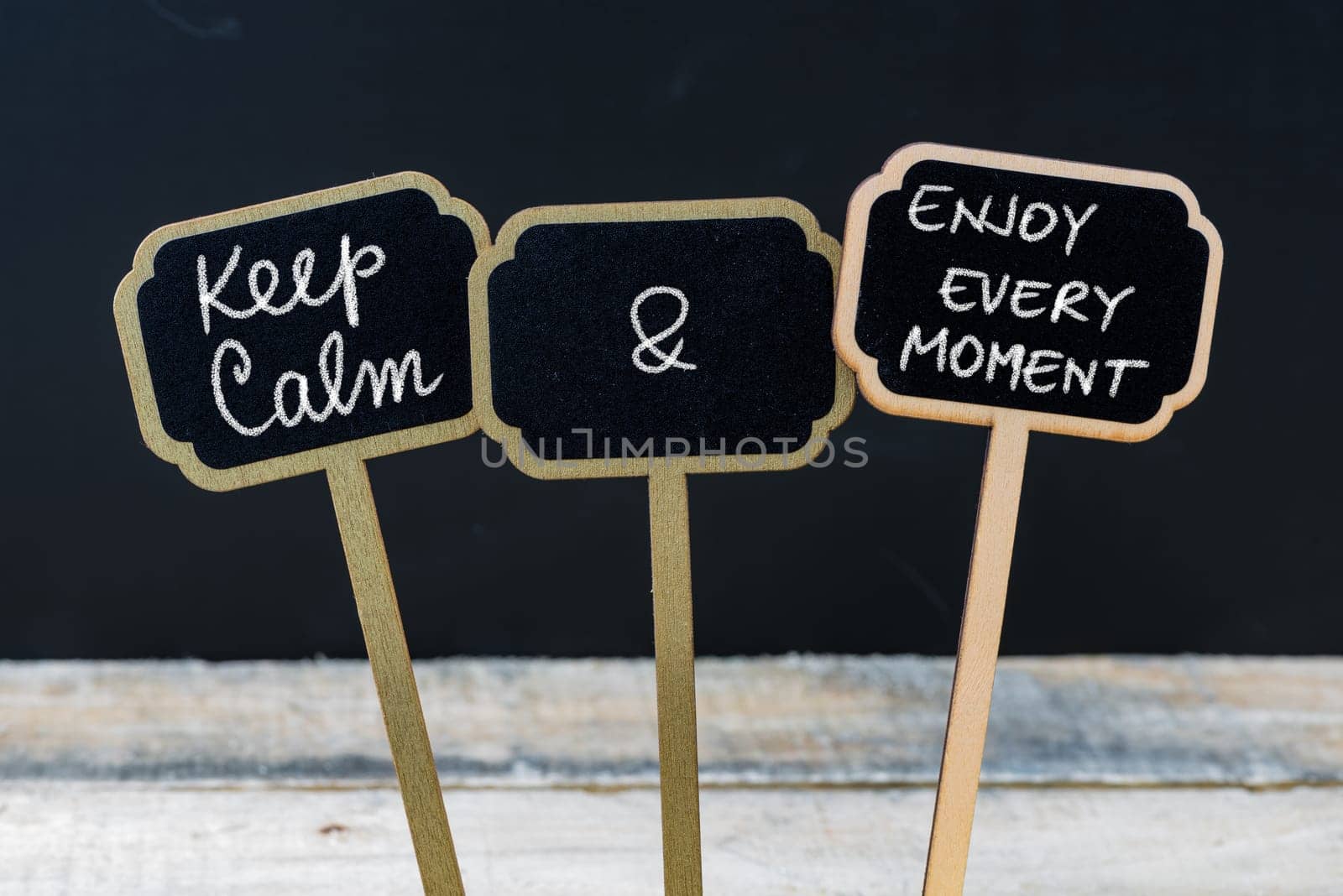 Keep Calm and Enjoy Every Moment message written with chalk on mini blackboard labels, defocused chalkboard and wooden table in background. Fun and humor concept