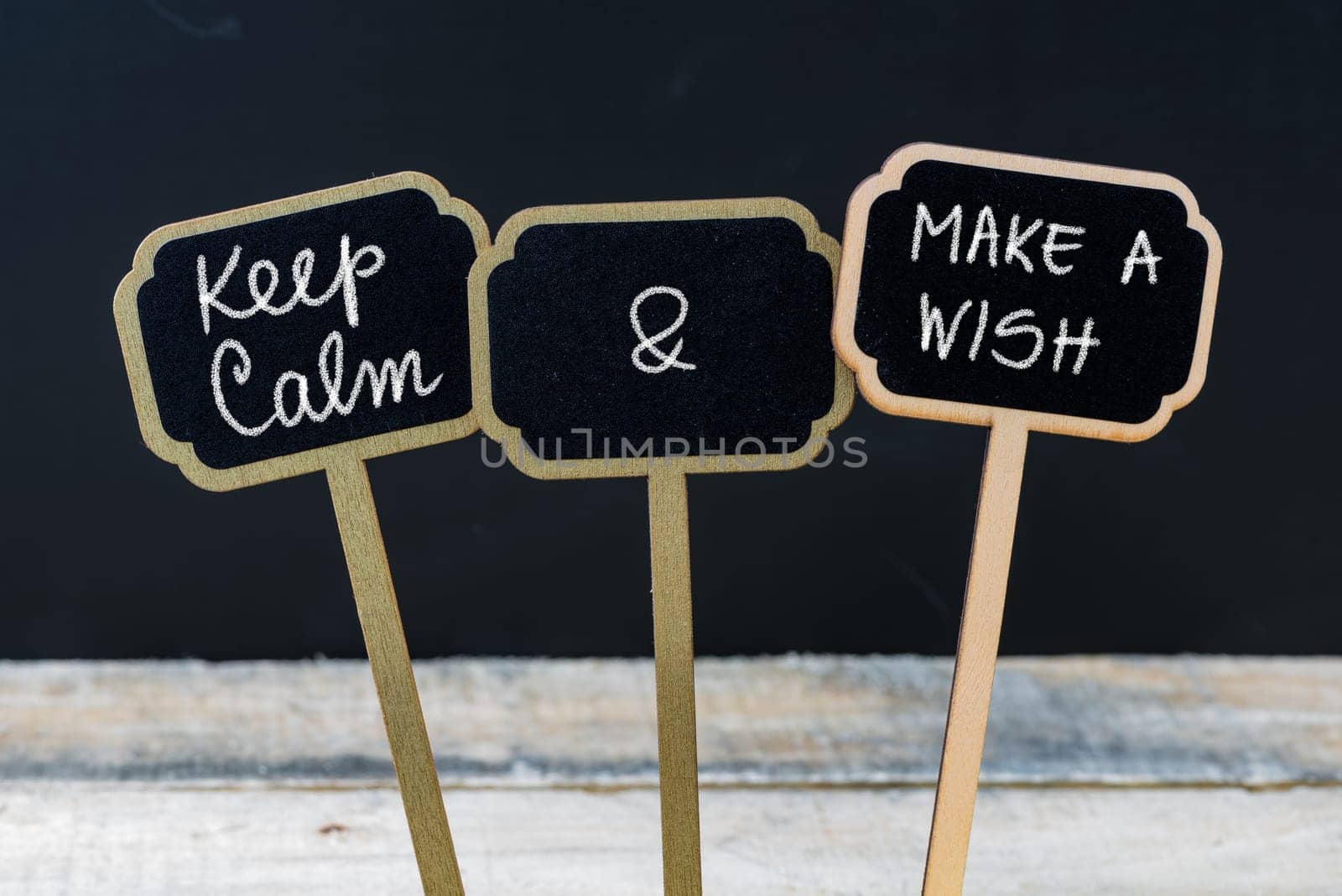 Keep Calm and Make A Wish message written with chalk on mini blackboard labels, defocused chalkboard and wooden table in background. Fun and humor concept