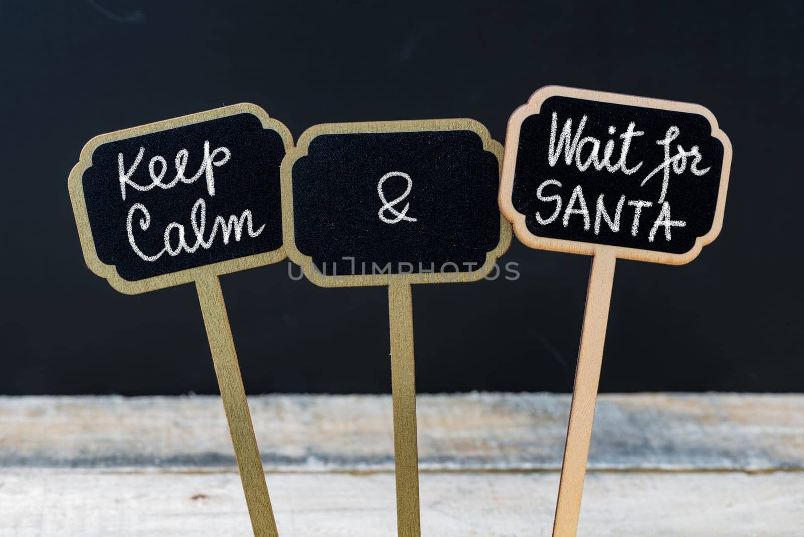 Keep Calm and Wait For Santa message written with chalk on mini blackboard labels, defocused chalkboard and wooden table in background. Fun and humor concept