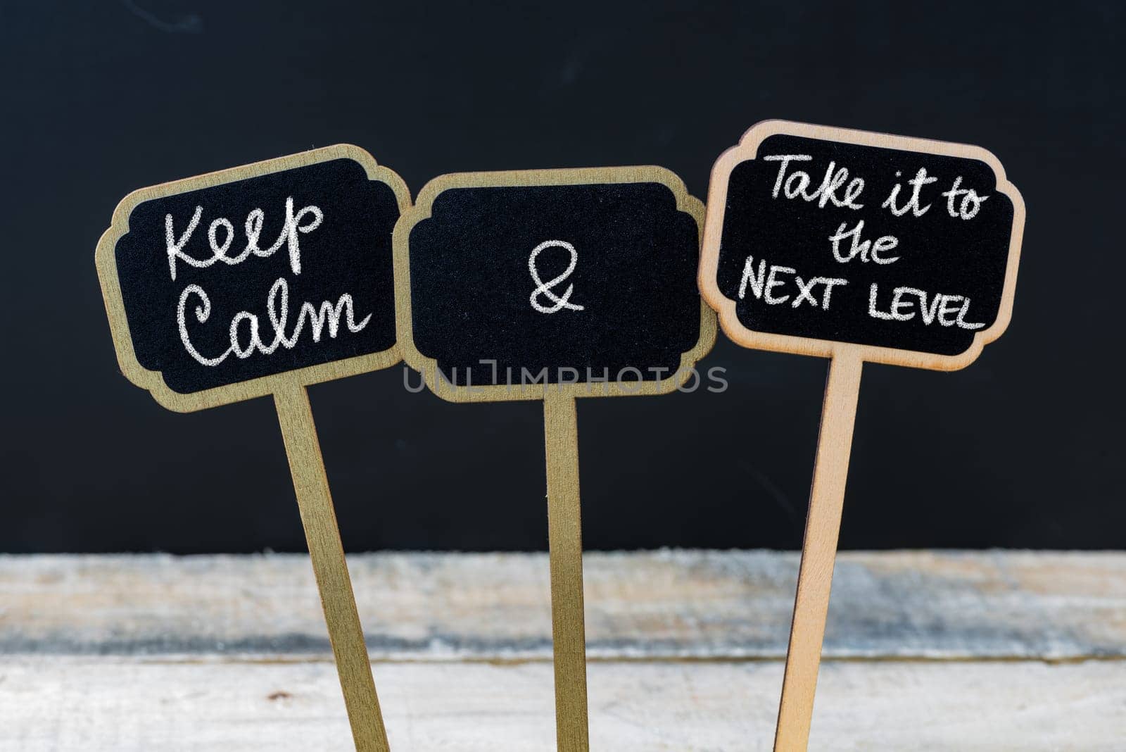 Keep Calm and Take It To The Next Level message written with chalk on mini blackboard labels, defocused chalkboard and wooden table in background. Fun and humor concept