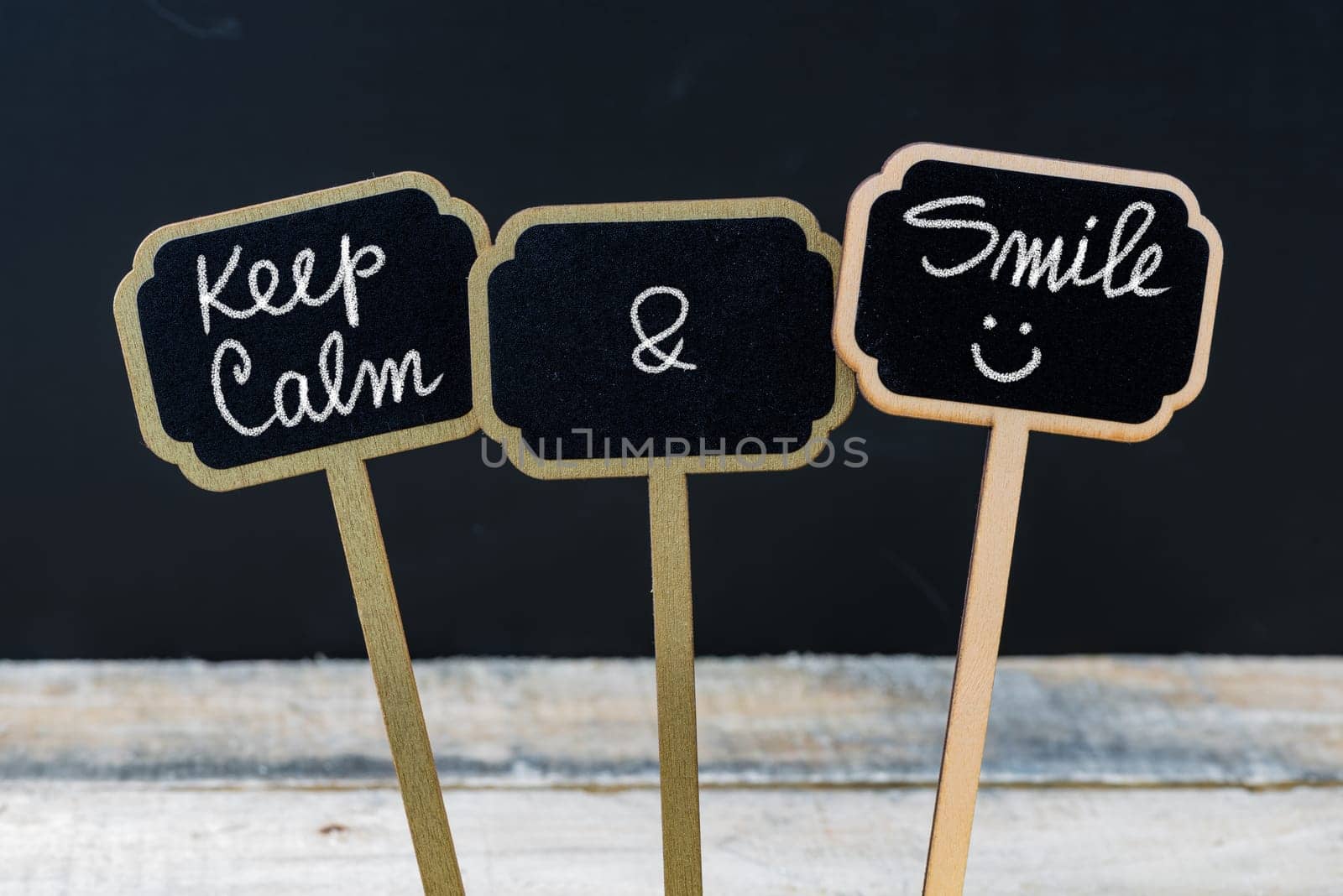 Keep Calm and Smile message written with chalk on mini blackboard labels, defocused chalkboard and wooden table in background. Fun and humor concept