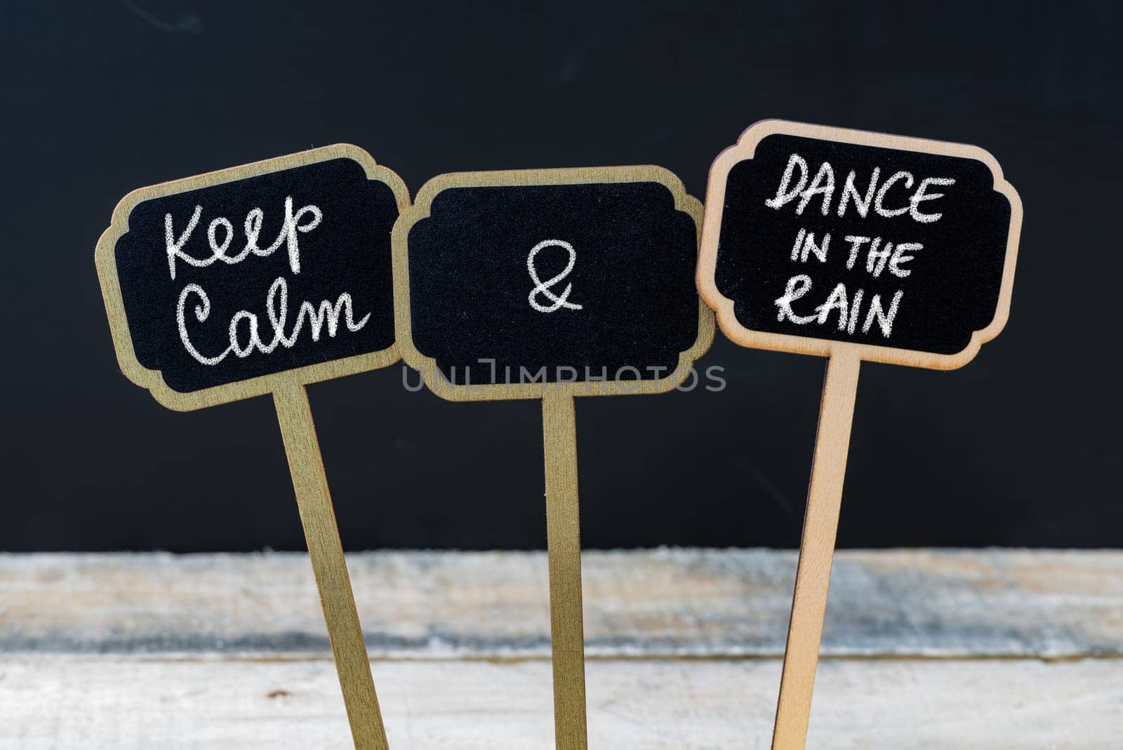 Keep Calm and Dance In The Rain message written with chalk on mini blackboard labels, defocused chalkboard and wooden table in background. Fun and humor concept