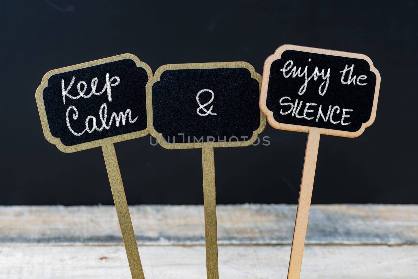 Keep Calm and Enjoy The Silence message written with chalk on mini blackboard labels, defocused chalkboard and wooden table in background. Fun and humor concept