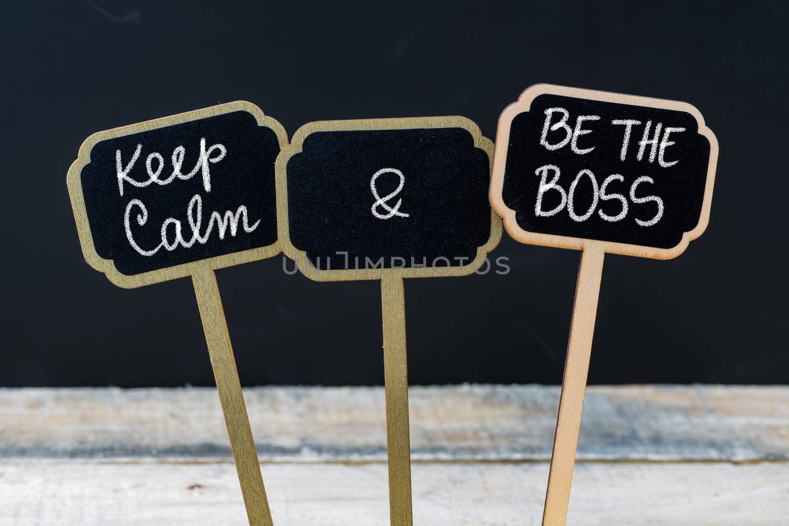 Keep Calm and Be The Boss message written with chalk on mini blackboard labels, defocused chalkboard and wooden table in background. Fun and humor concept