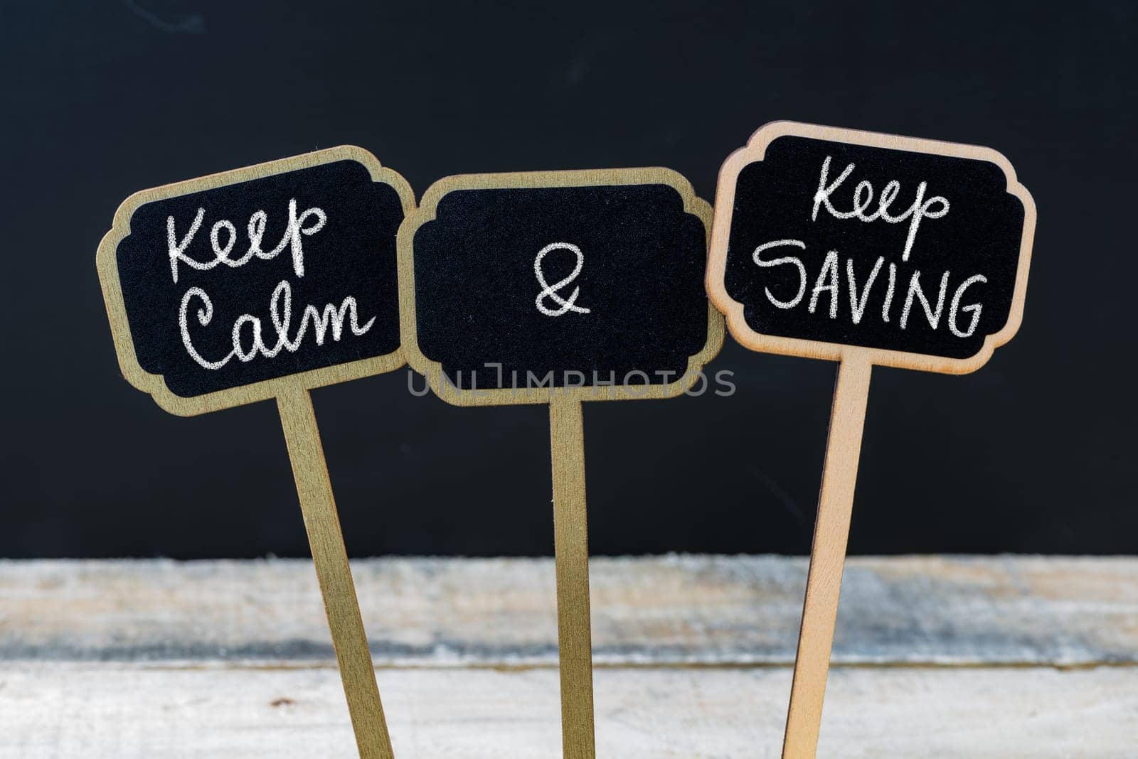 Keep Calm and Keep Saving message written with chalk on mini blackboard labels, defocused chalkboard and wooden table in background. Fun and humor concept