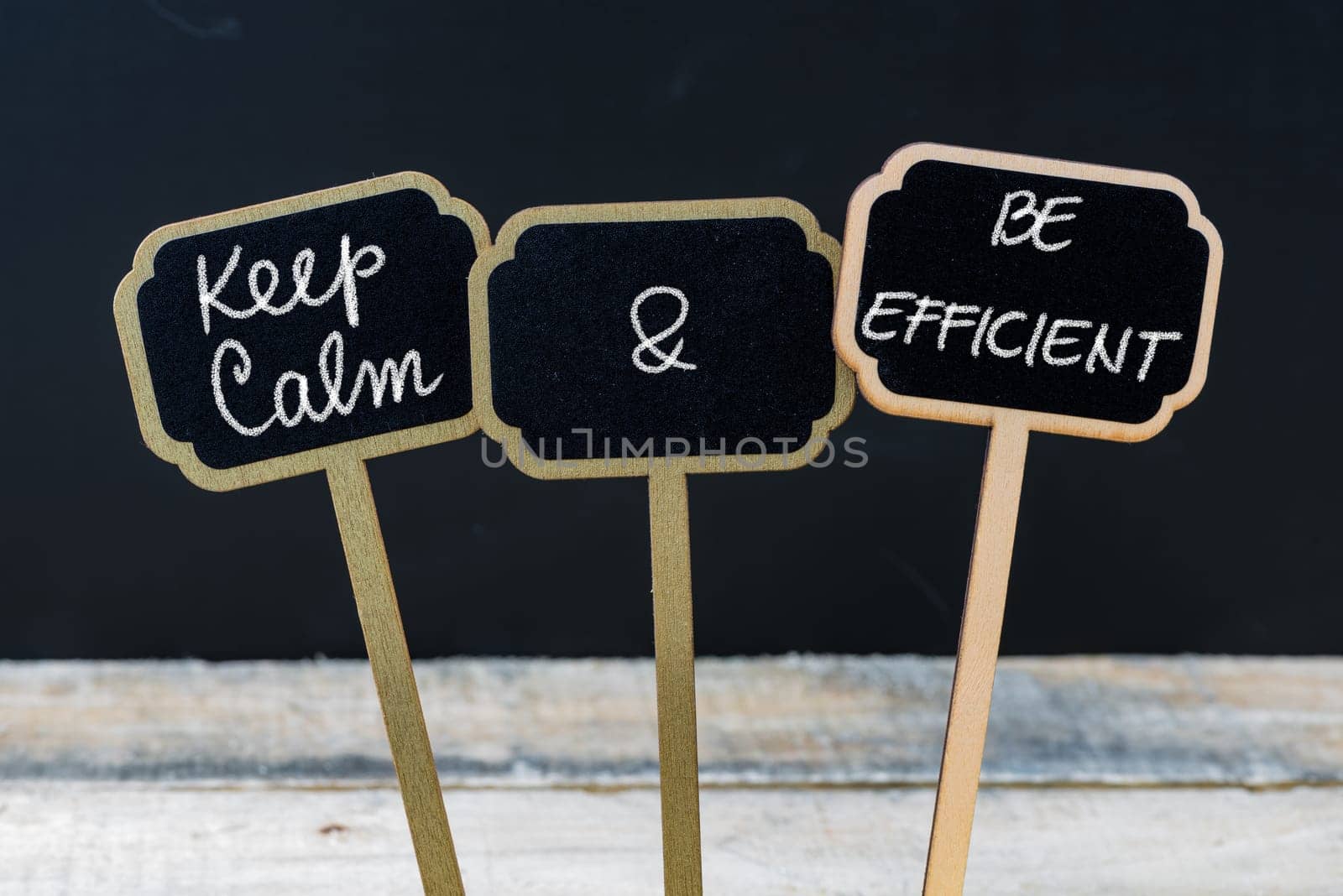Keep Calm and Be Efficient message written with chalk on mini blackboard labels, defocused chalkboard and wooden table in background. Fun and humor concept