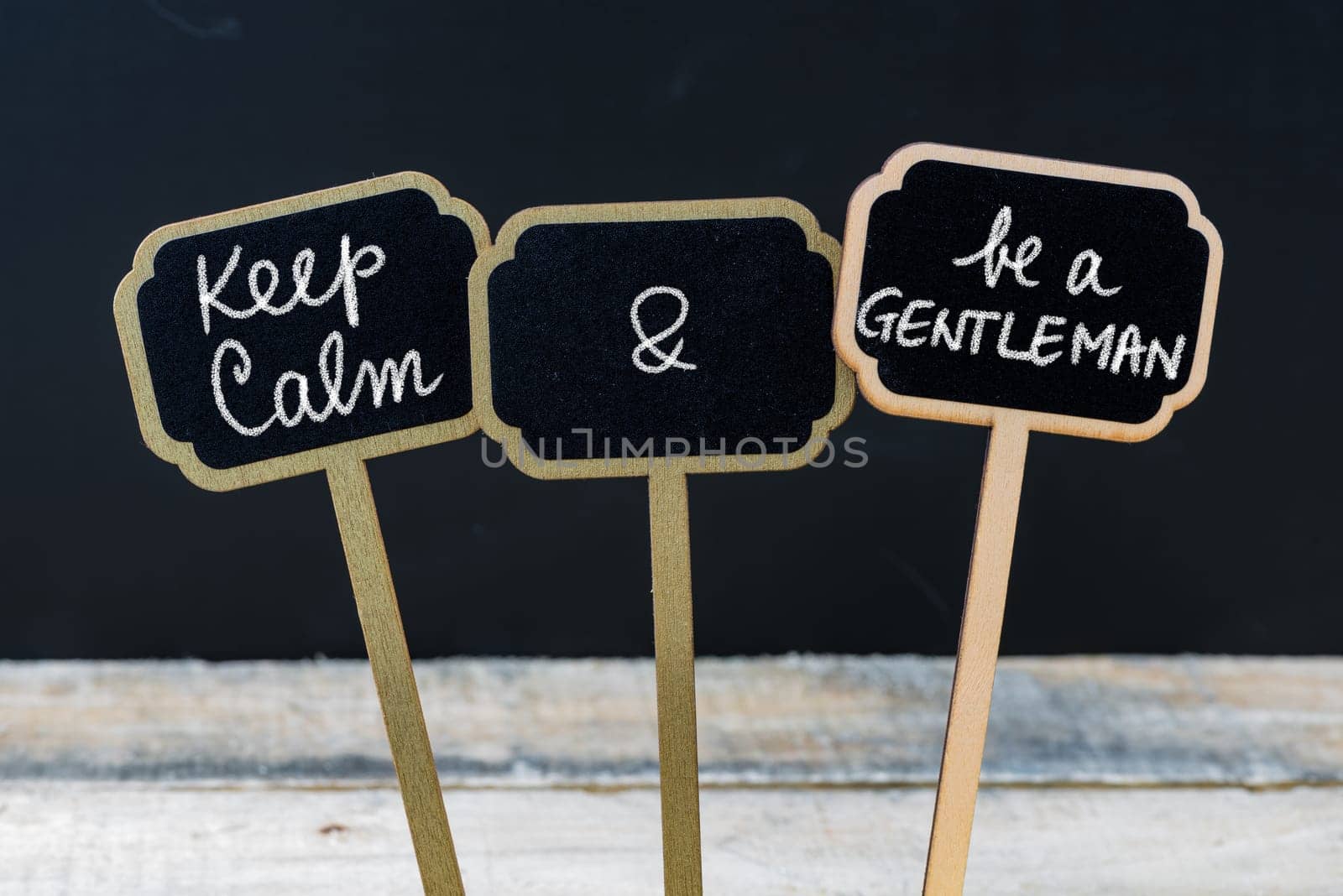 Keep Calm and Be A Gentleman message written with chalk on mini blackboard labels, defocused chalkboard and wooden table in background. Fun and humor concept