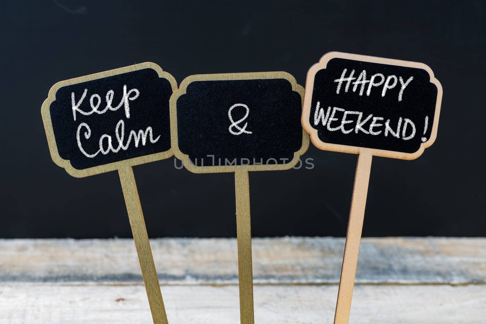 Keep Calm and Happy Weekend message written with chalk on mini blackboard labels, defocused chalkboard and wooden table in background. Fun and humor concept