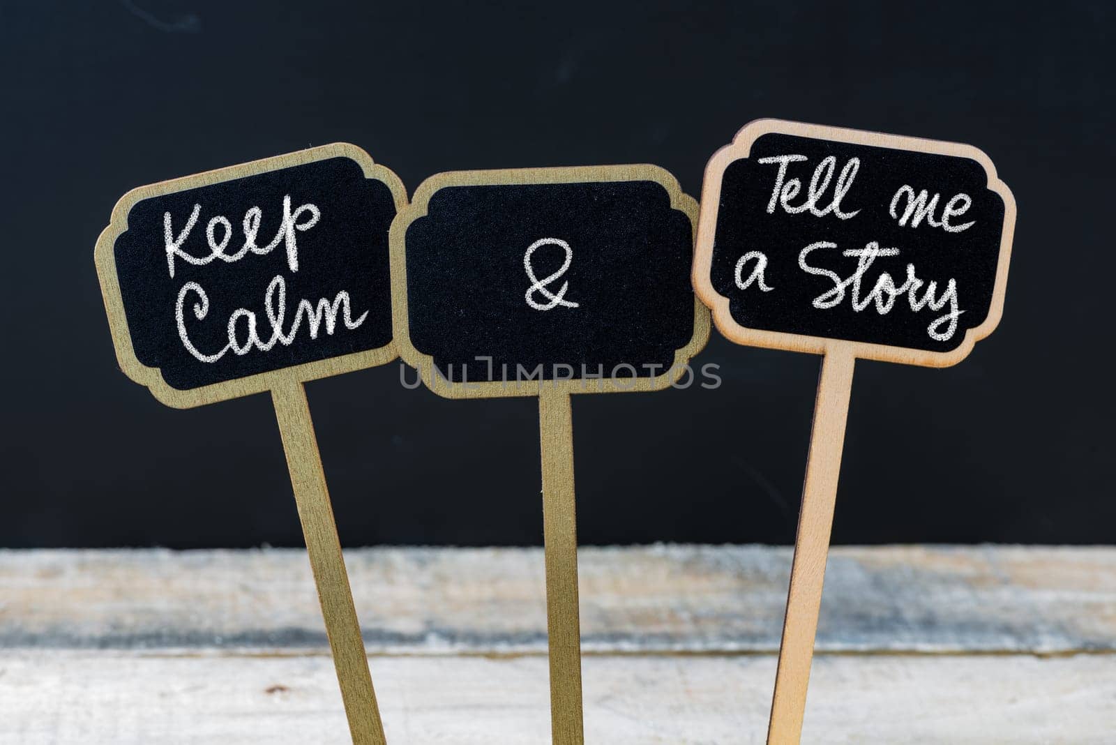 Keep Calm and Tell Me A Story message written with chalk on mini blackboard labels, defocused chalkboard and wooden table in background. Fun and humor concept