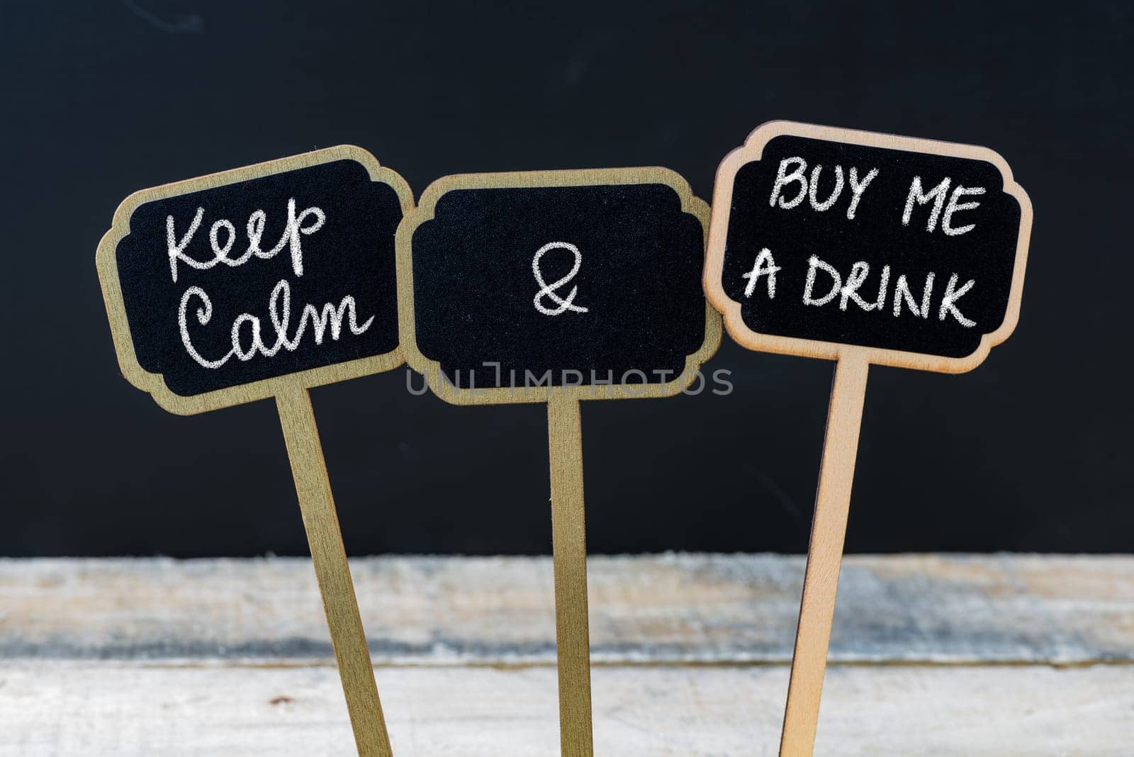 Keep Calm and Buy Me A Drink message written with chalk on mini blackboard labels, defocused chalkboard and wooden table in background. Fun and humor concept