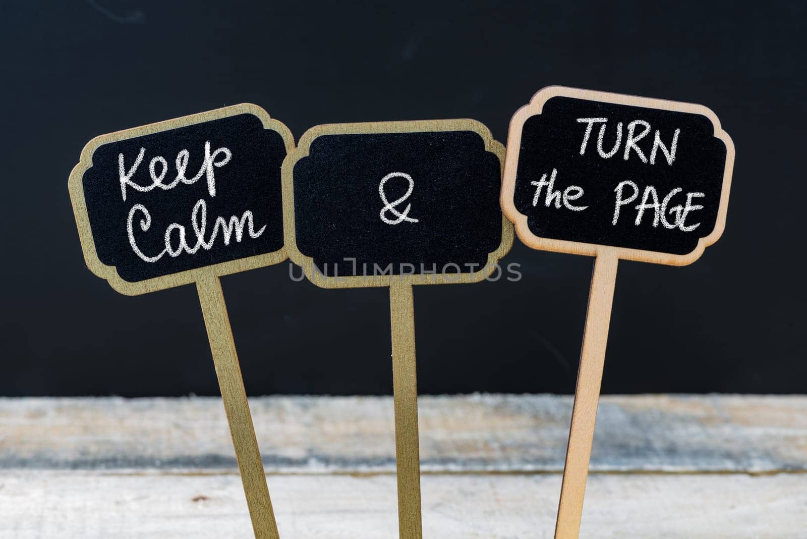 Keep Calm and Turn The Page message written with chalk on mini blackboard labels, defocused chalkboard and wooden table in background. Fun and humor concept