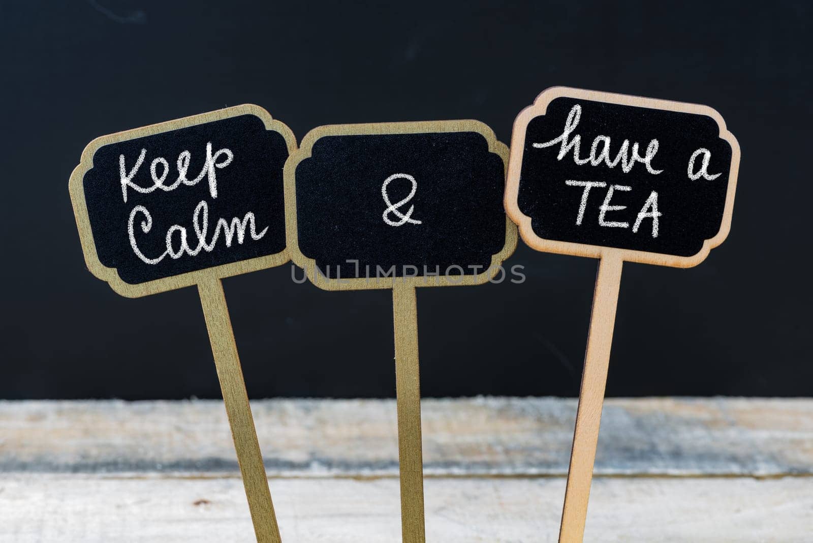 Keep Calm and Have A Tea message written with chalk on mini blackboard labels, defocused chalkboard and wooden table in background. Fun and humor concept